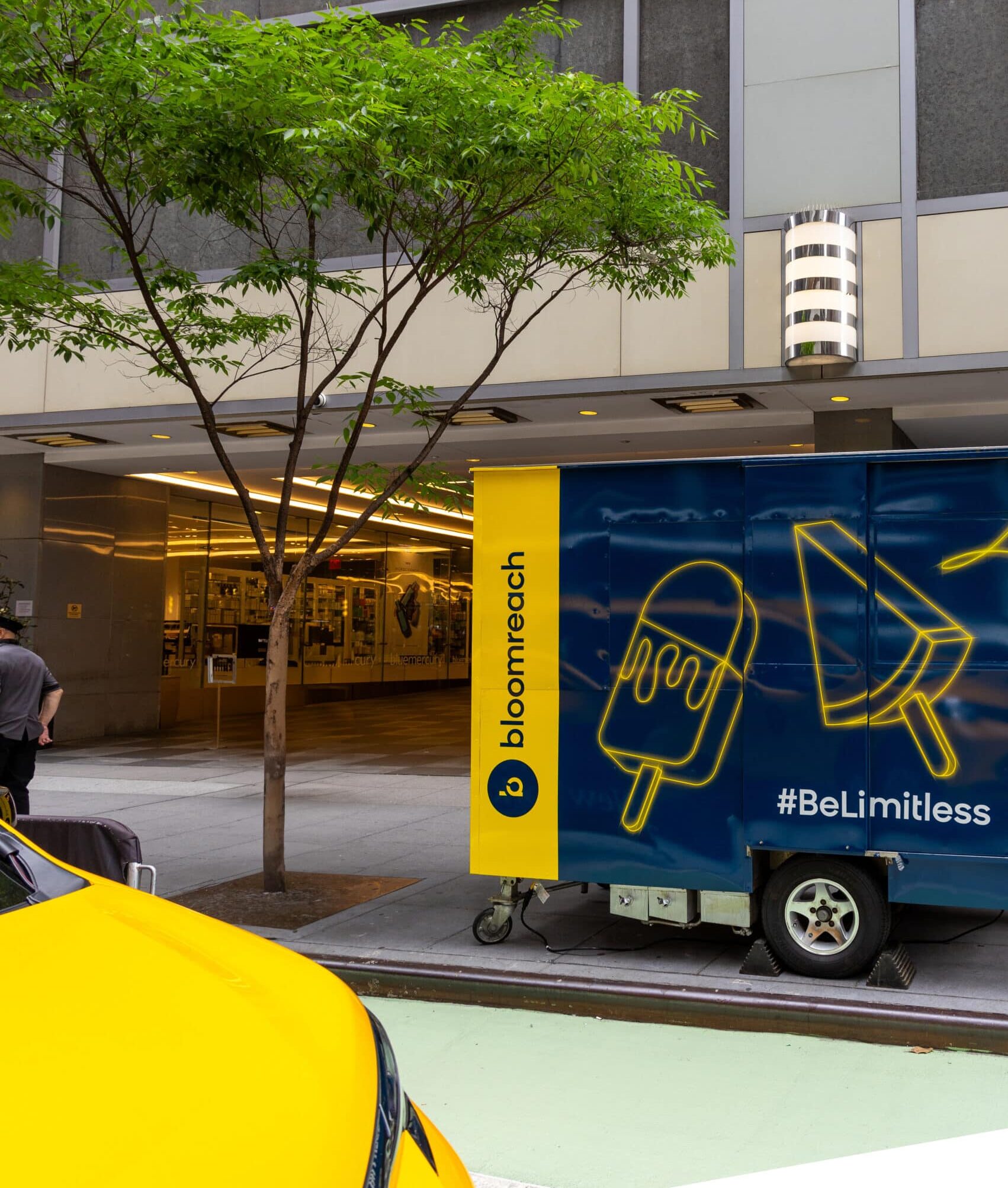 A blue Bloomreach popsicle truck is parked by a tree on a city street near a yellow car.