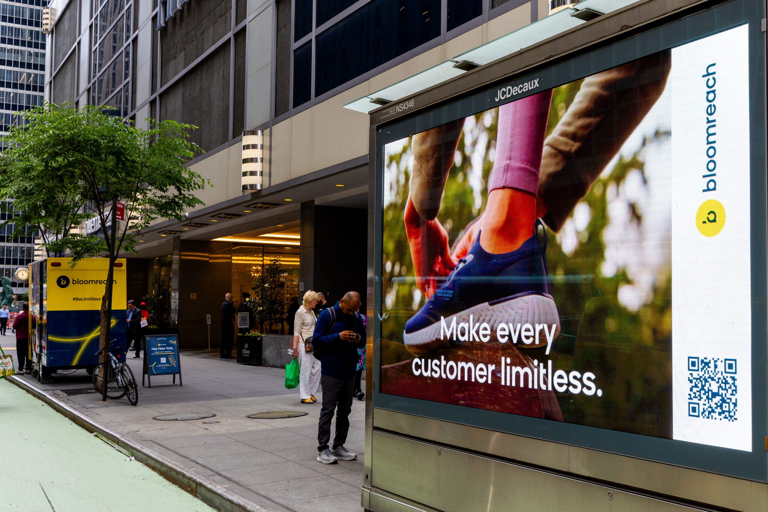 Digital billboard shows runners' shoes ad; people walk by offices, popsicle stand, and another ad.