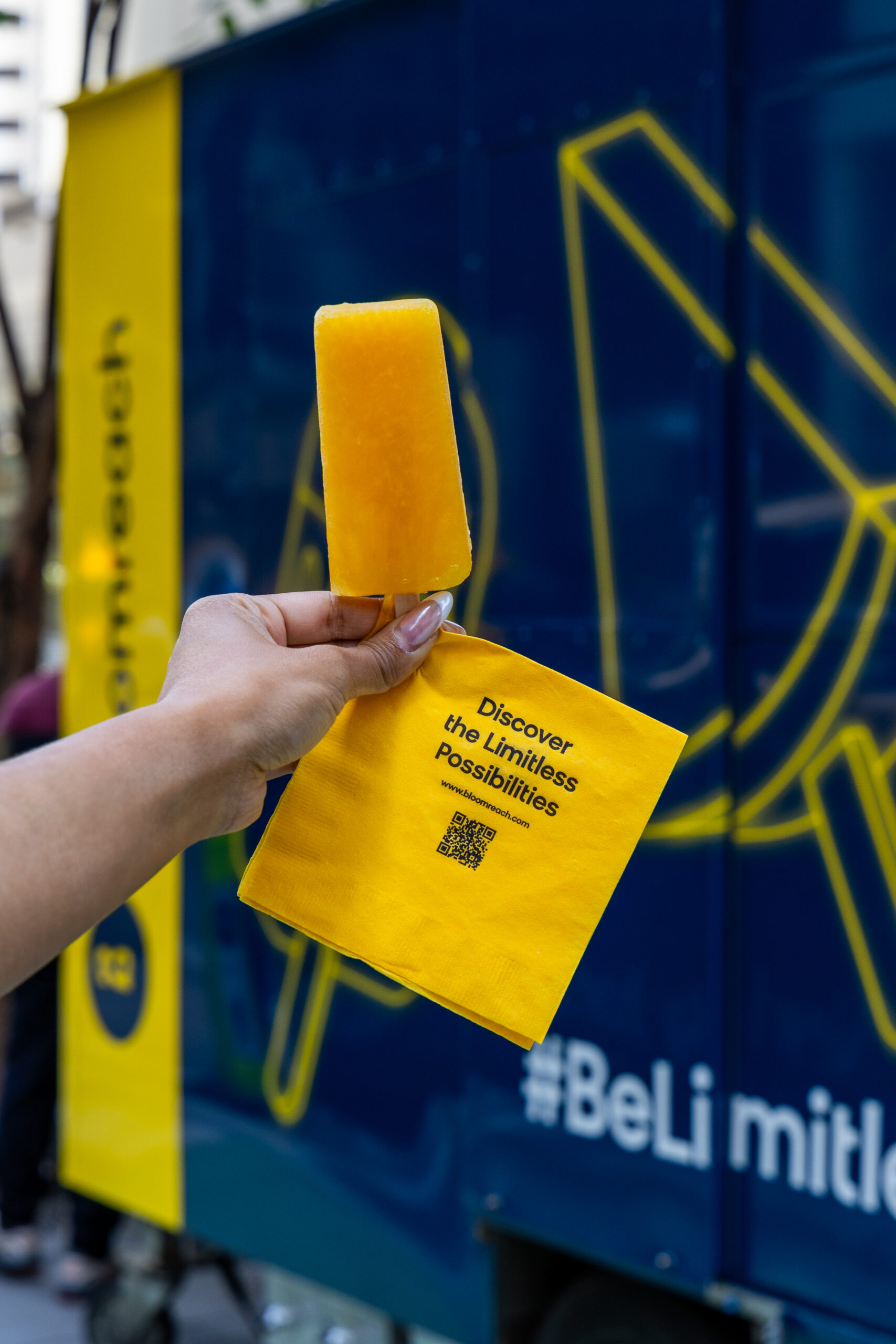 Hand holding a yellow popsicle and napkin with QR code in front of a blue and yellow popsicle truck.