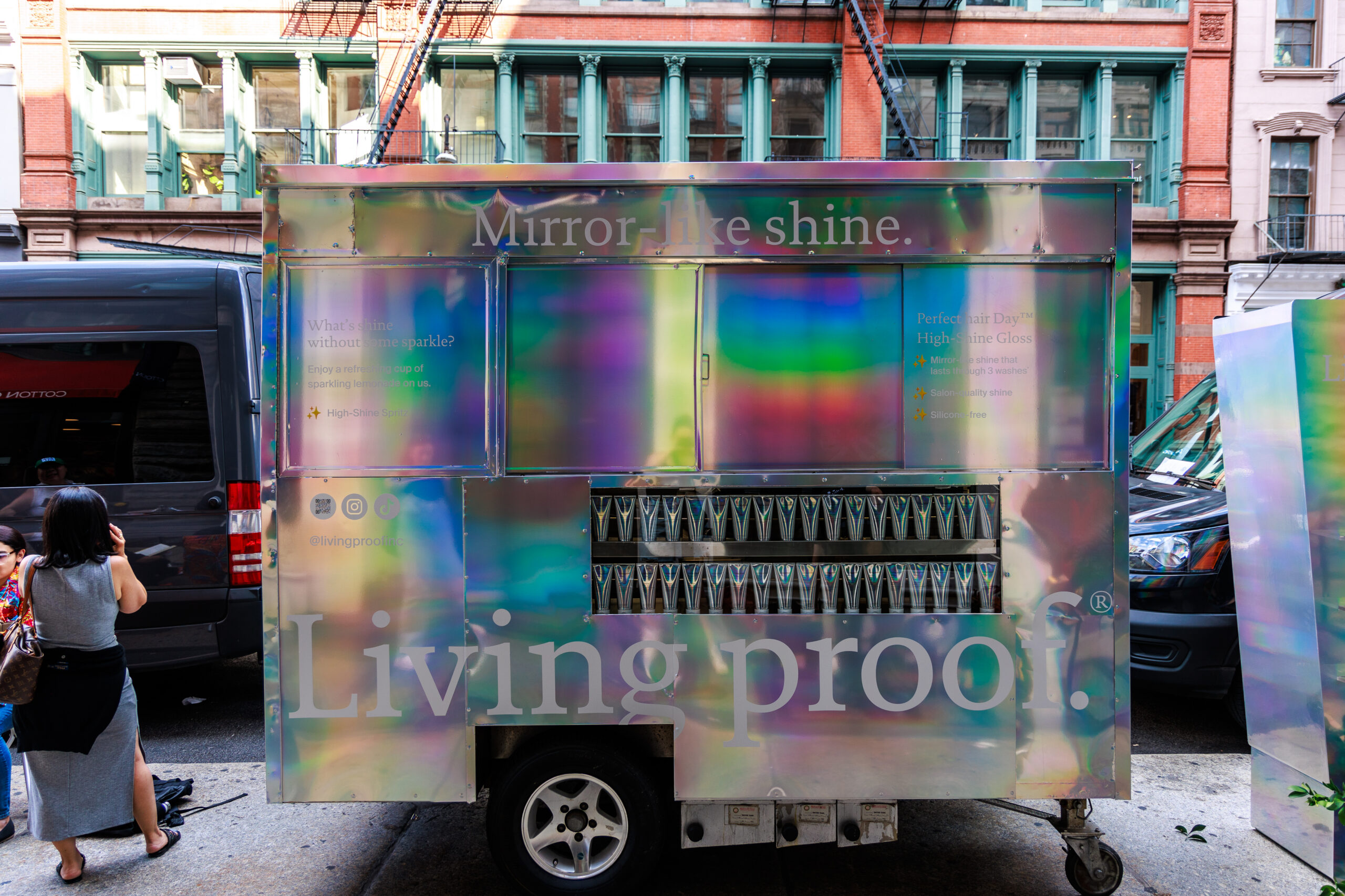 A Living Proof promo truck with hair products is parked on a city street; a woman takes a photo.