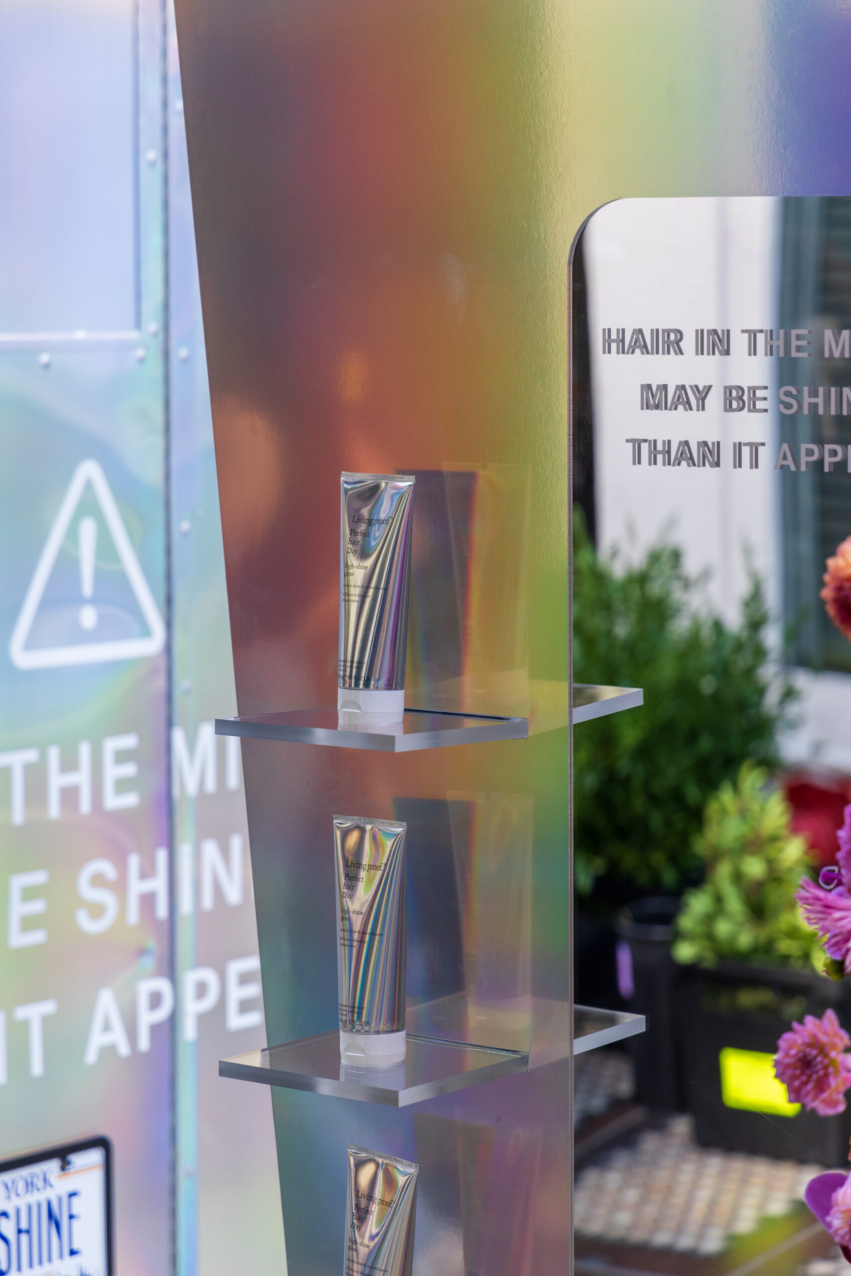 Three rainbow-striped hair product tubes on a shelf with flowers and a partial sign behind.