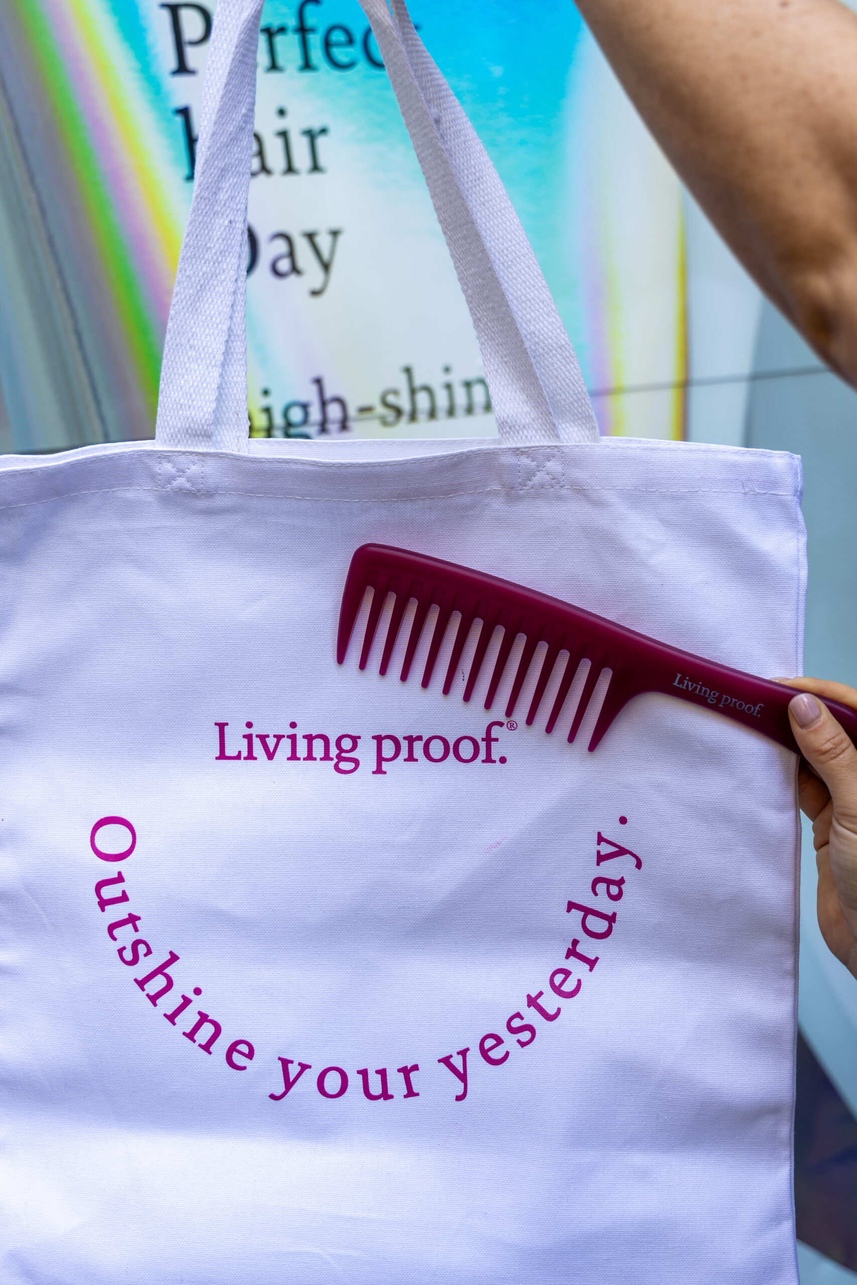 Hand holding maroon comb over white tote bag with pink text; blurred sign in background.