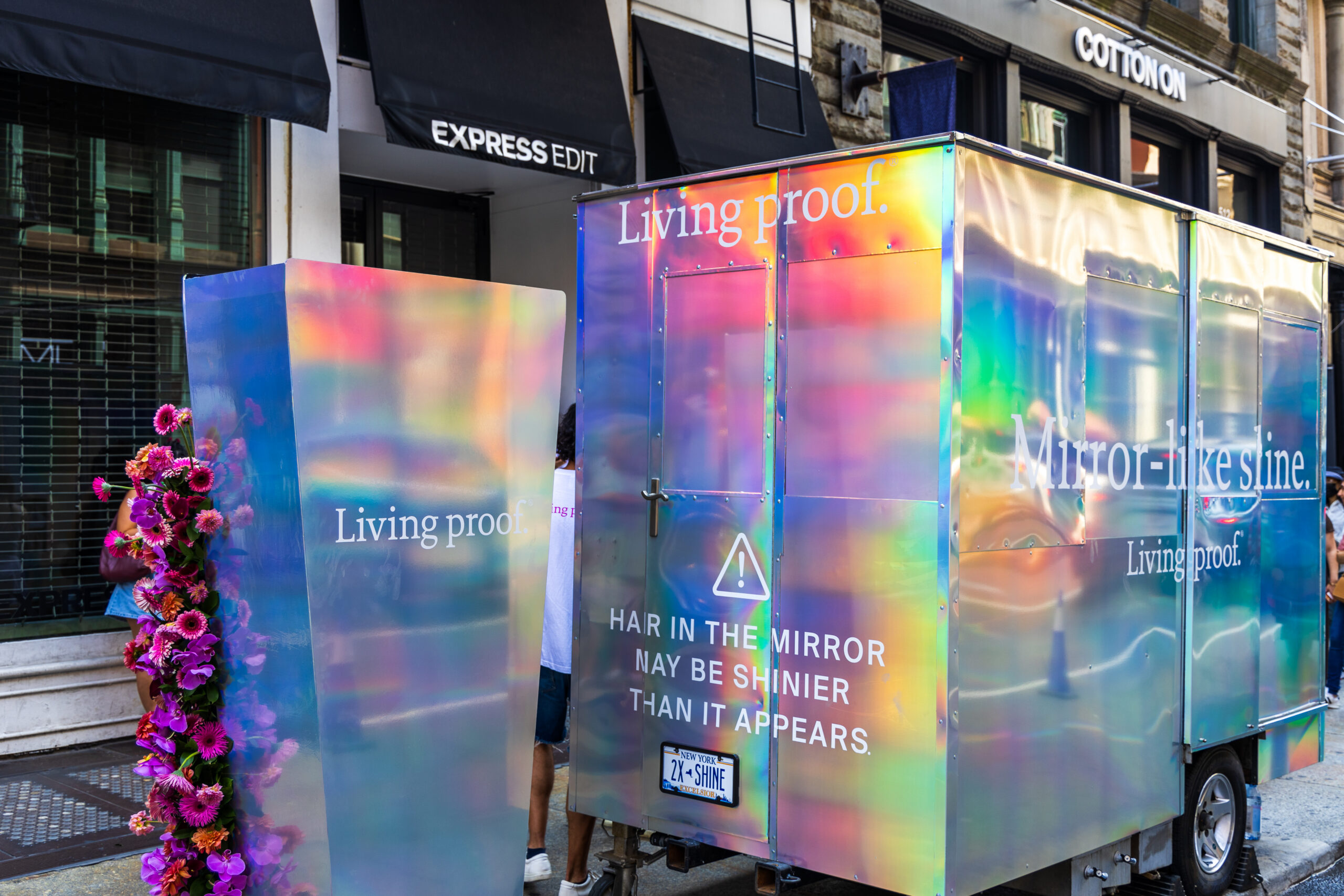 A shiny popsicle trailer with hair shine slogans is parked in the city; pink flowers nearby.