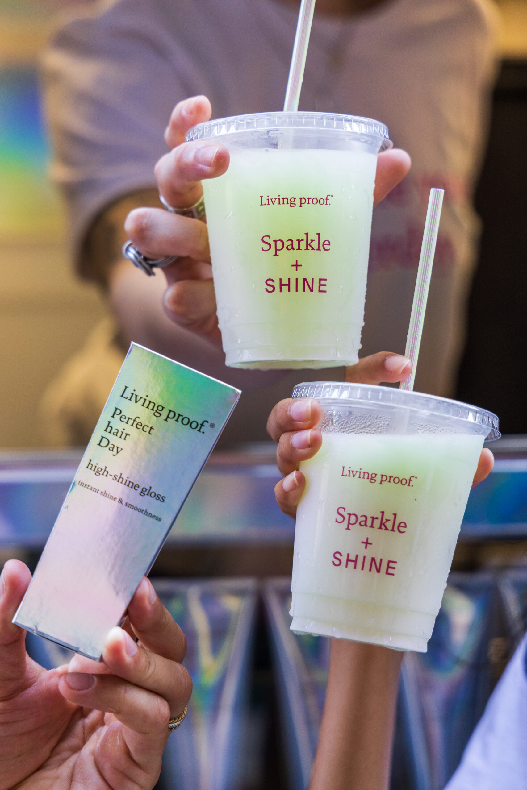 Three hands hold two cups with green drinks and a box, all labeled Living proof, at a pop-up.