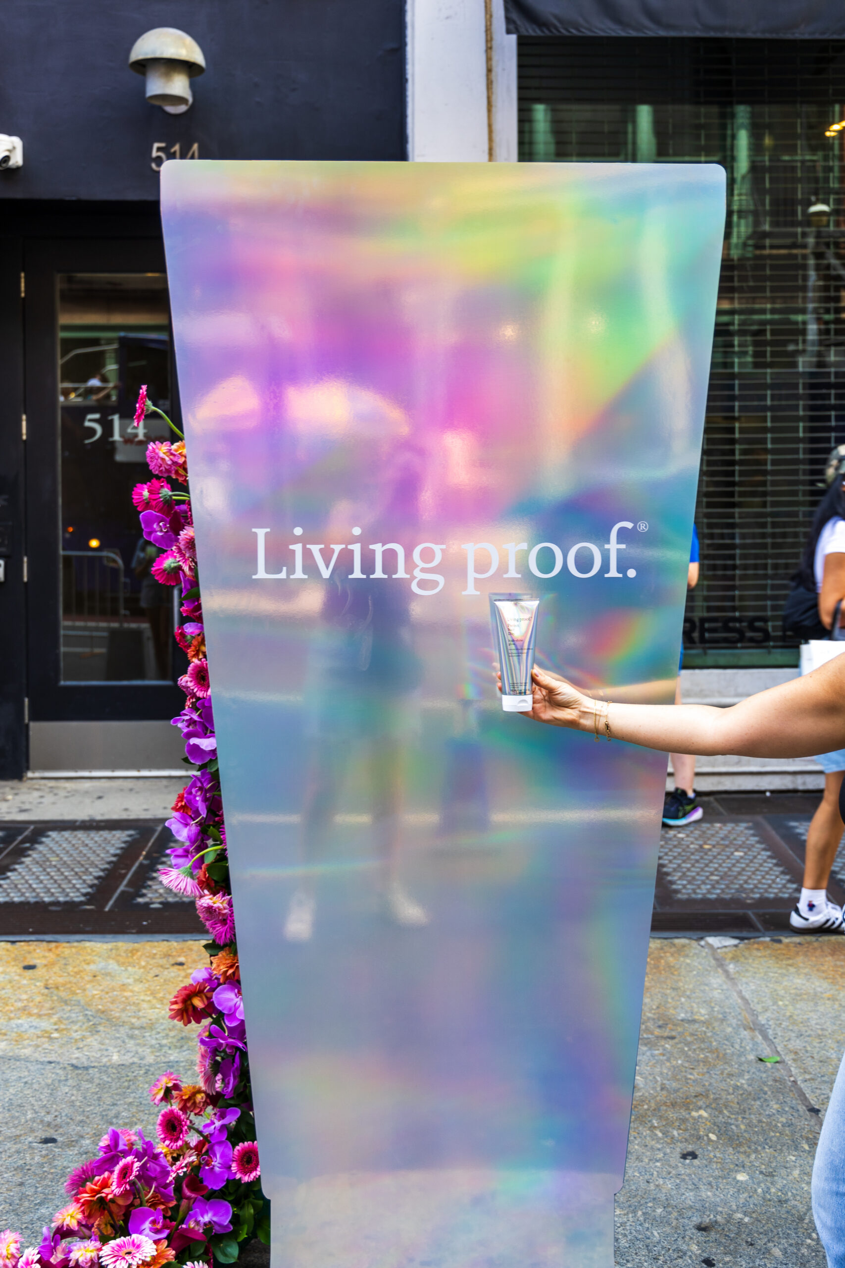 Hand holding glass near “Living proof.” sign, with purple and pink flowers on city sidewalk.