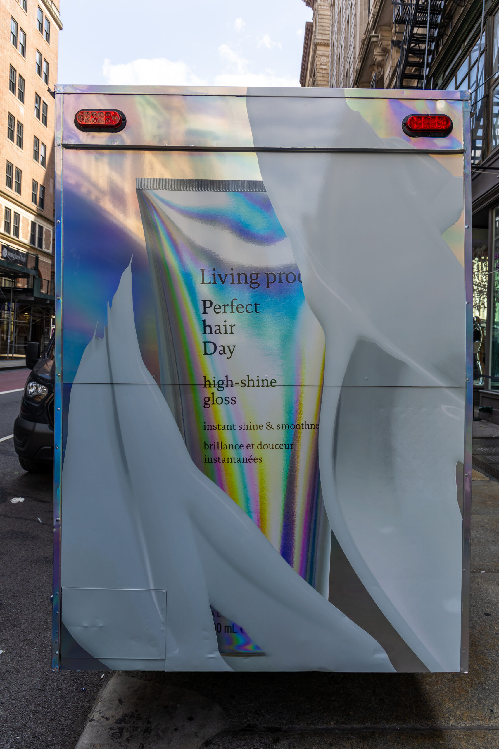 Truck with ad for Living proof high-shine gloss, parked on city street near popsicle stand.