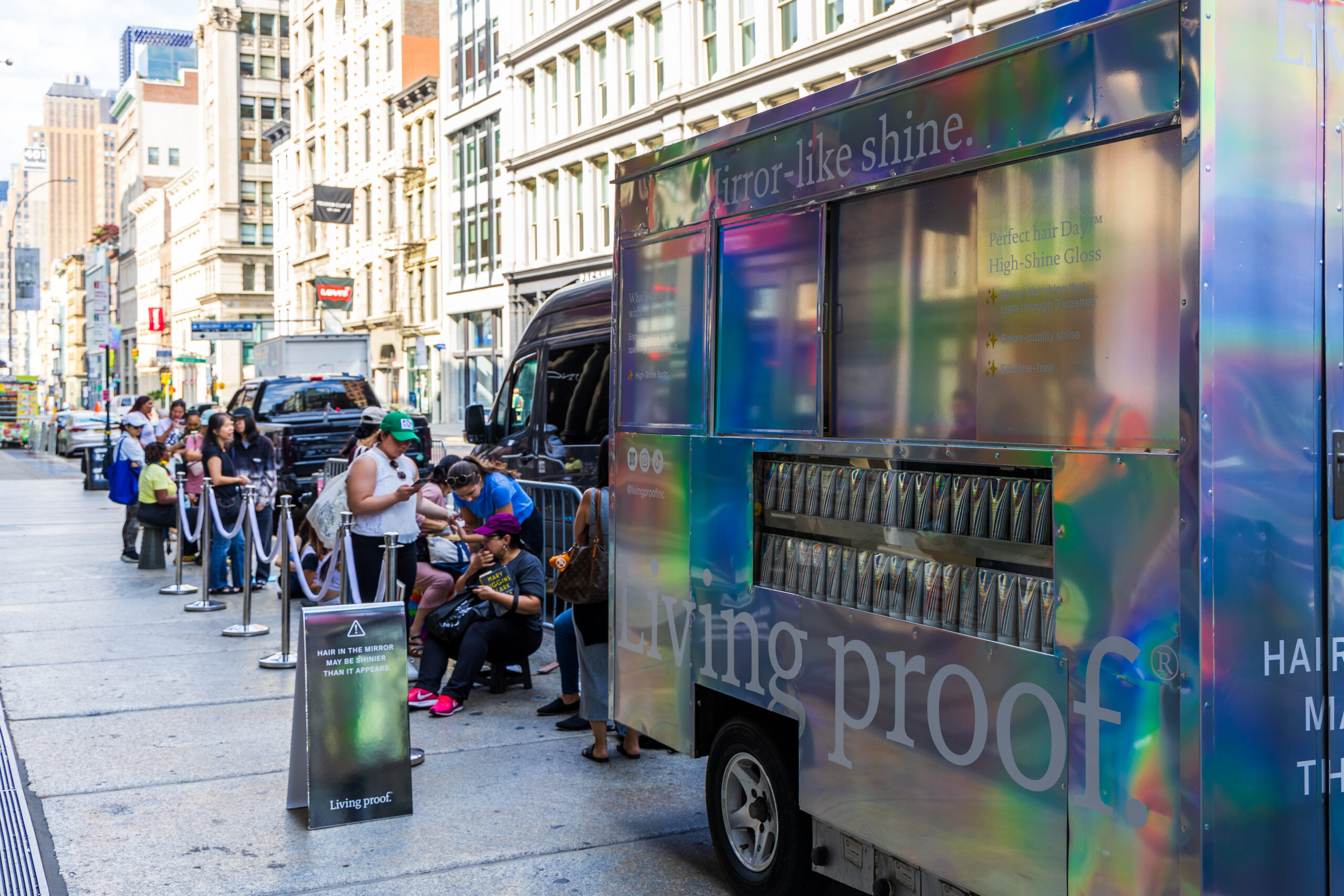 People line up outside a Living Proof popsicle pop-up truck on a city sidewalk with tall buildings.