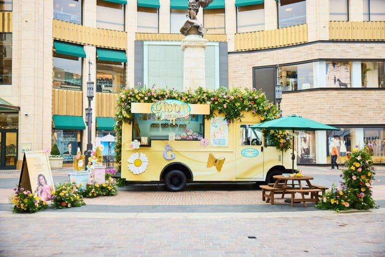 Pastel yellow ice cream truck with flowers and butterflies parked in a plaza by big buildings.