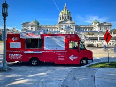 Red pizza food truck parked by large government building with green dome and clear blue sky.
