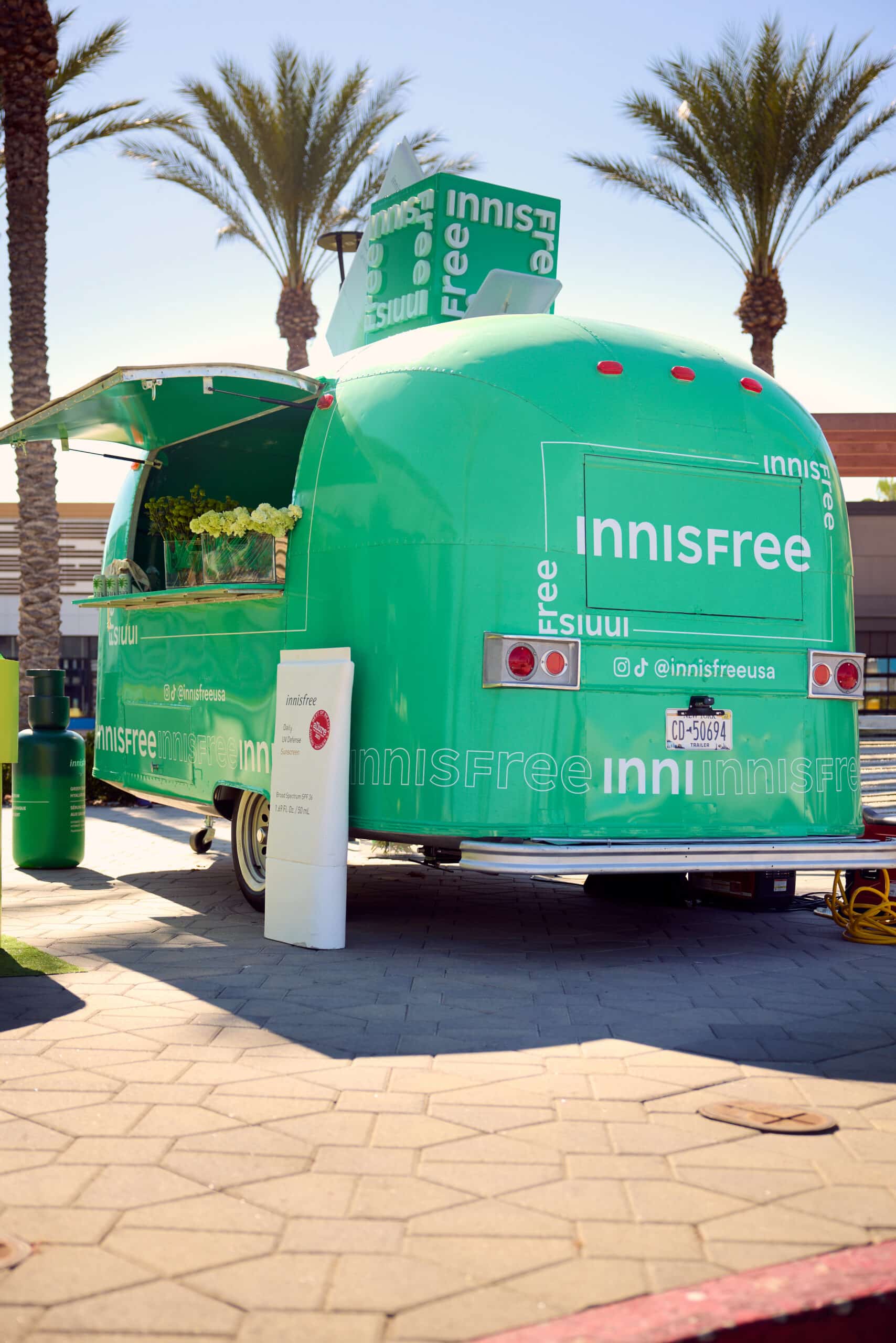 Green Innisfree trailer with open hatch parked under palm trees, logo and products visible.
