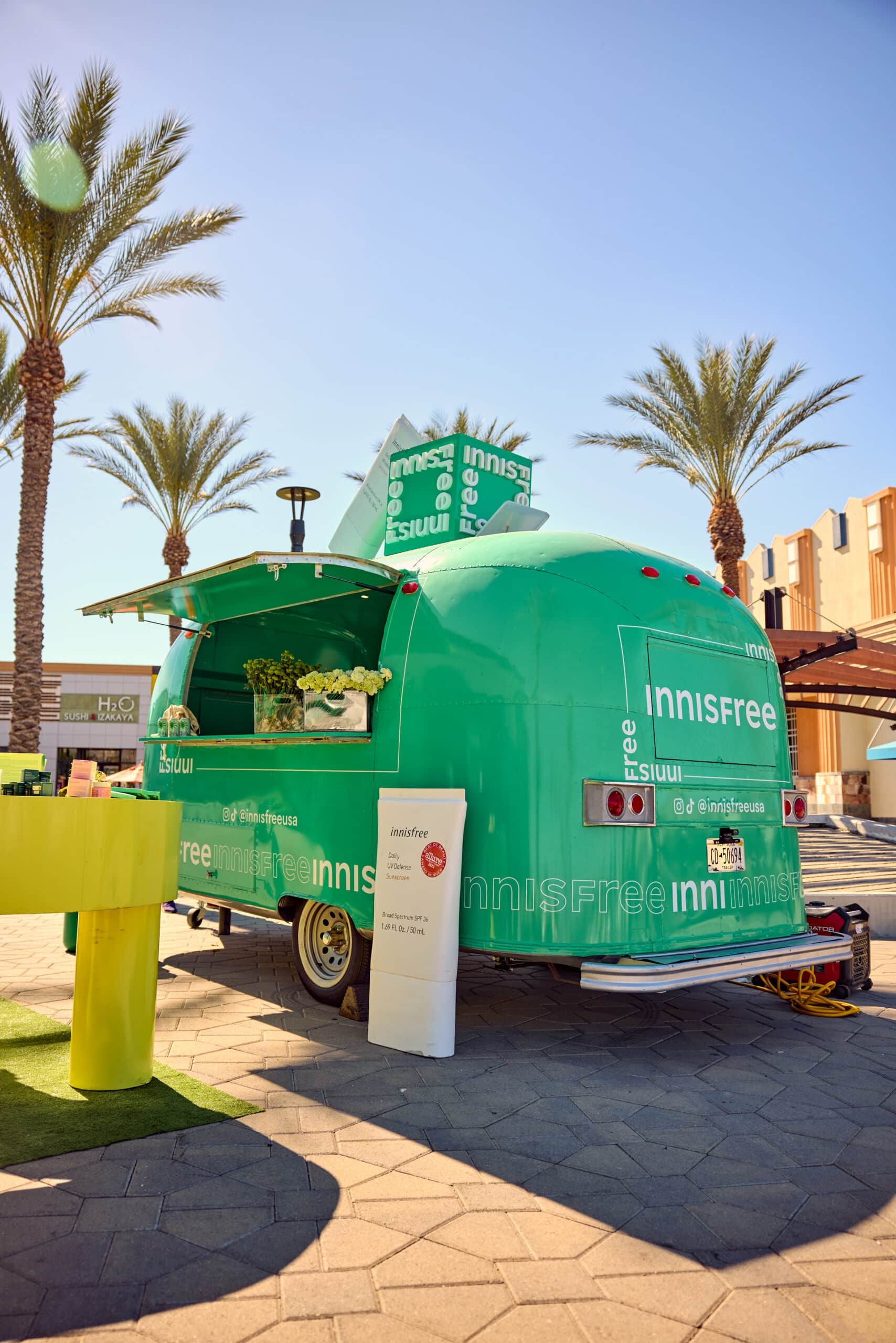 Green Innisfree airstream trailer with open window parked outside near palm trees and tables.