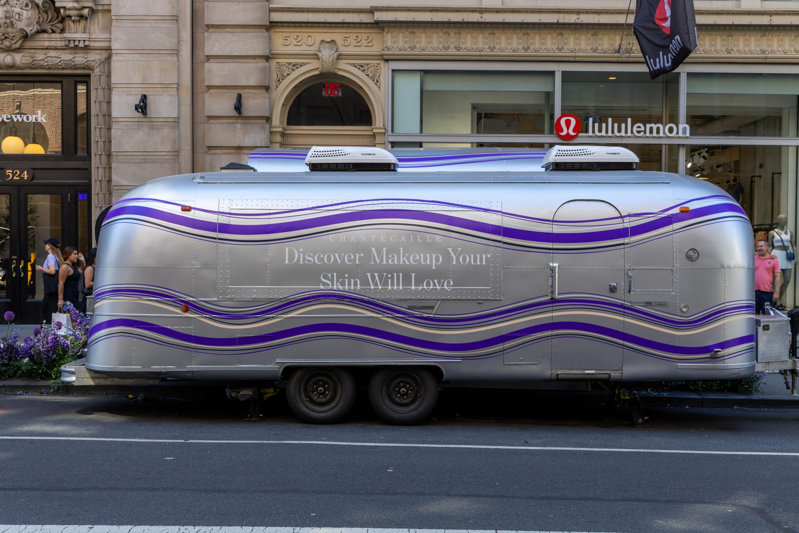 A silver trailer with purple designs and makeup ad text is parked outside a Lululemon store.