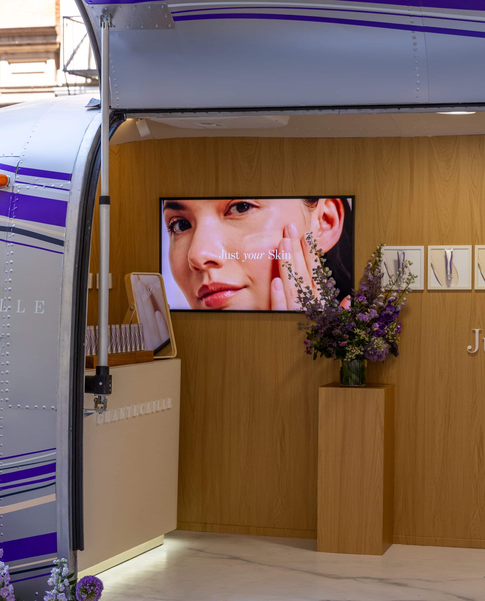 A screen in an airstream shows a woman and "Just your Skin"; purple flowers in vase nearby.