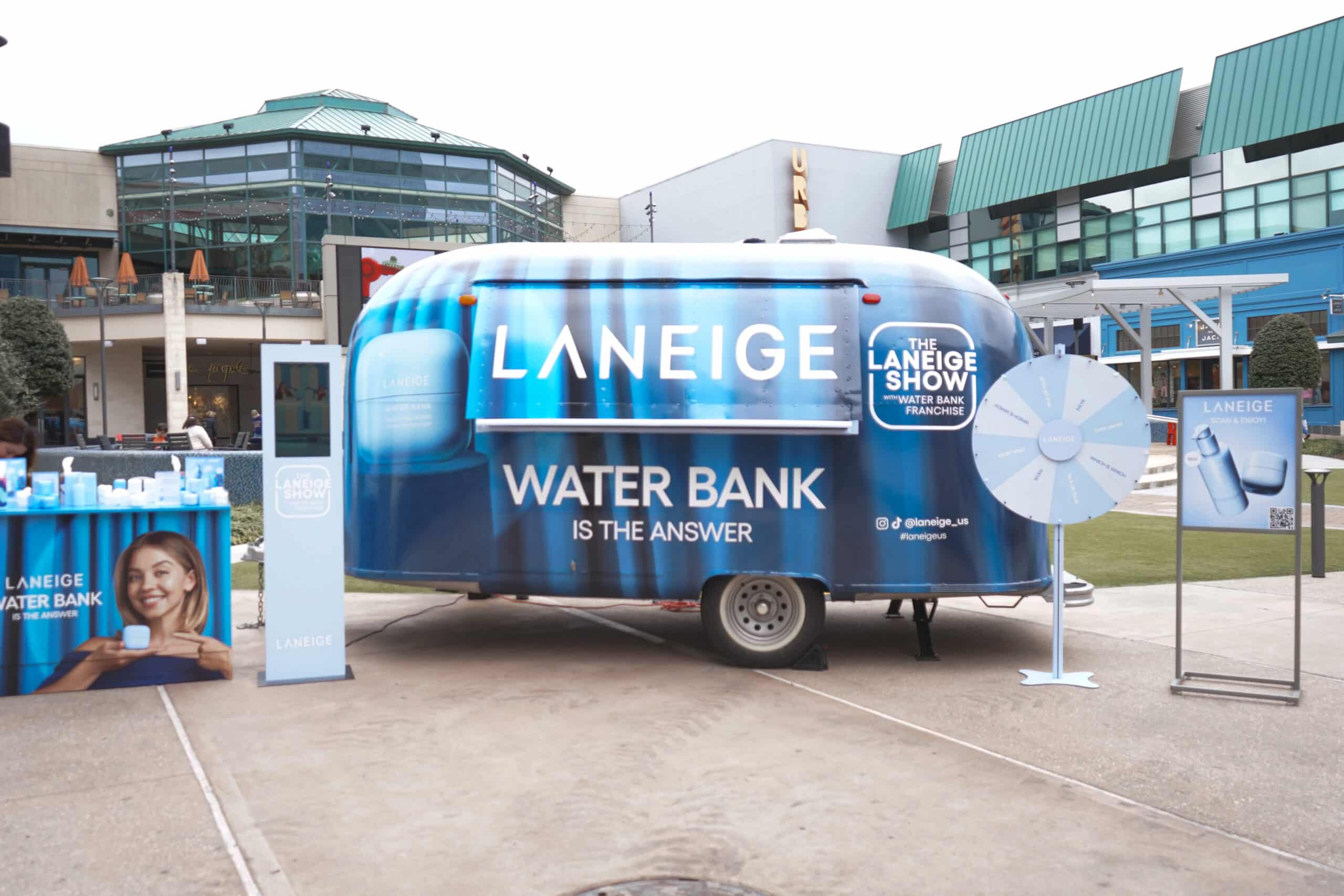 Blue Laneige airstream trailer with promo stand, spinning wheel, and products in shopping area.