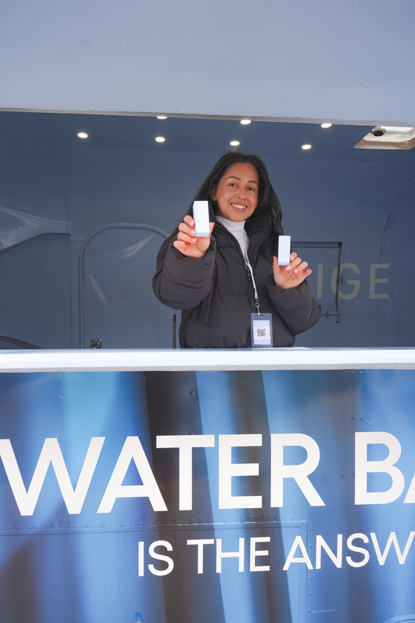 Person in a black jacket stands behind a WATER BAR sign, holding two small white objects.