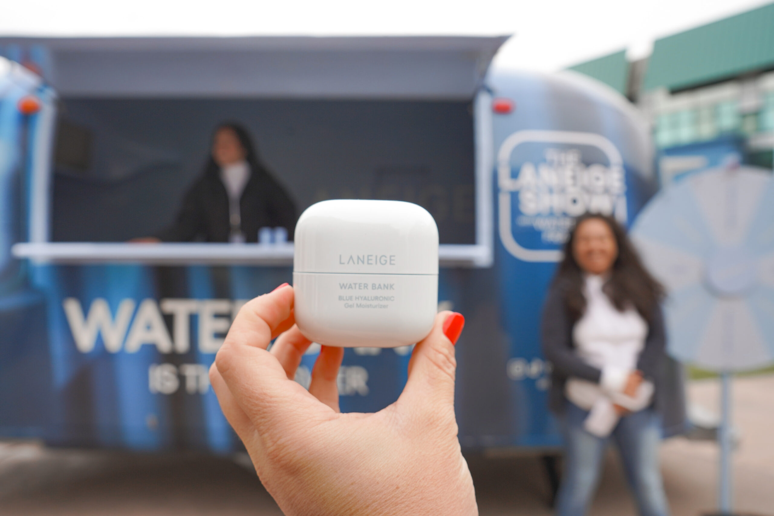 Hand holds Laneige Water Bank moisturizer; blue booth and two people blurred in background.