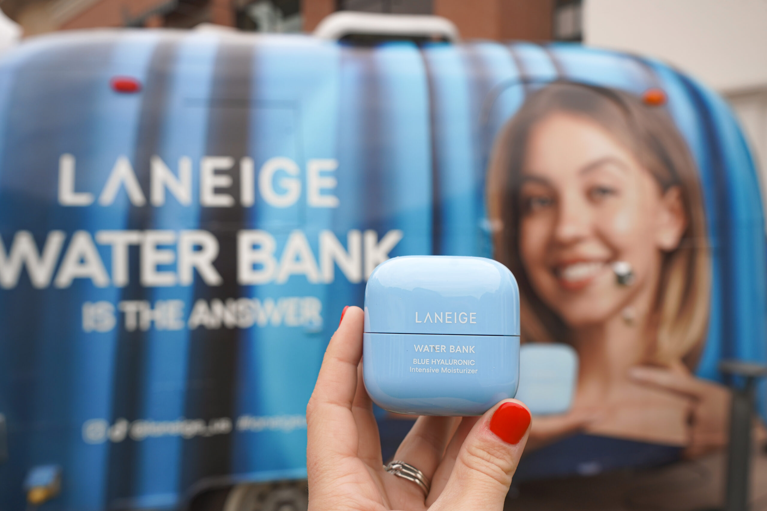 Hand with red nails holds blue Laneige moisturizer in front of trailer with Laneige ad and woman.