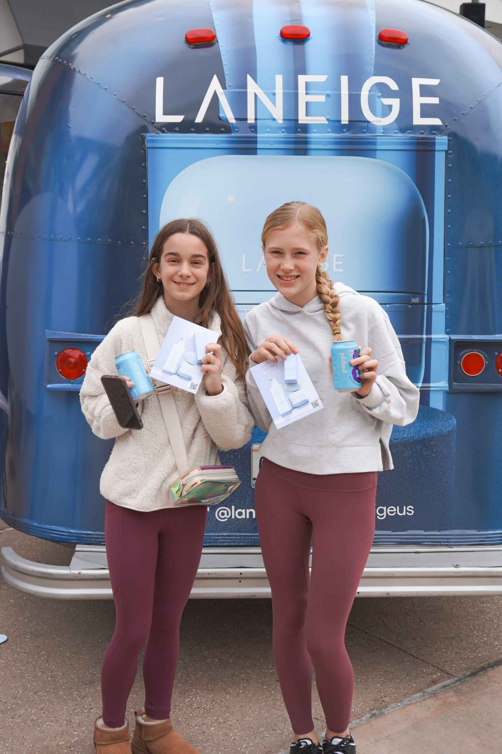 Two girls in maroon leggings and white tops hold Laneige samples and cups in front of a trailer.