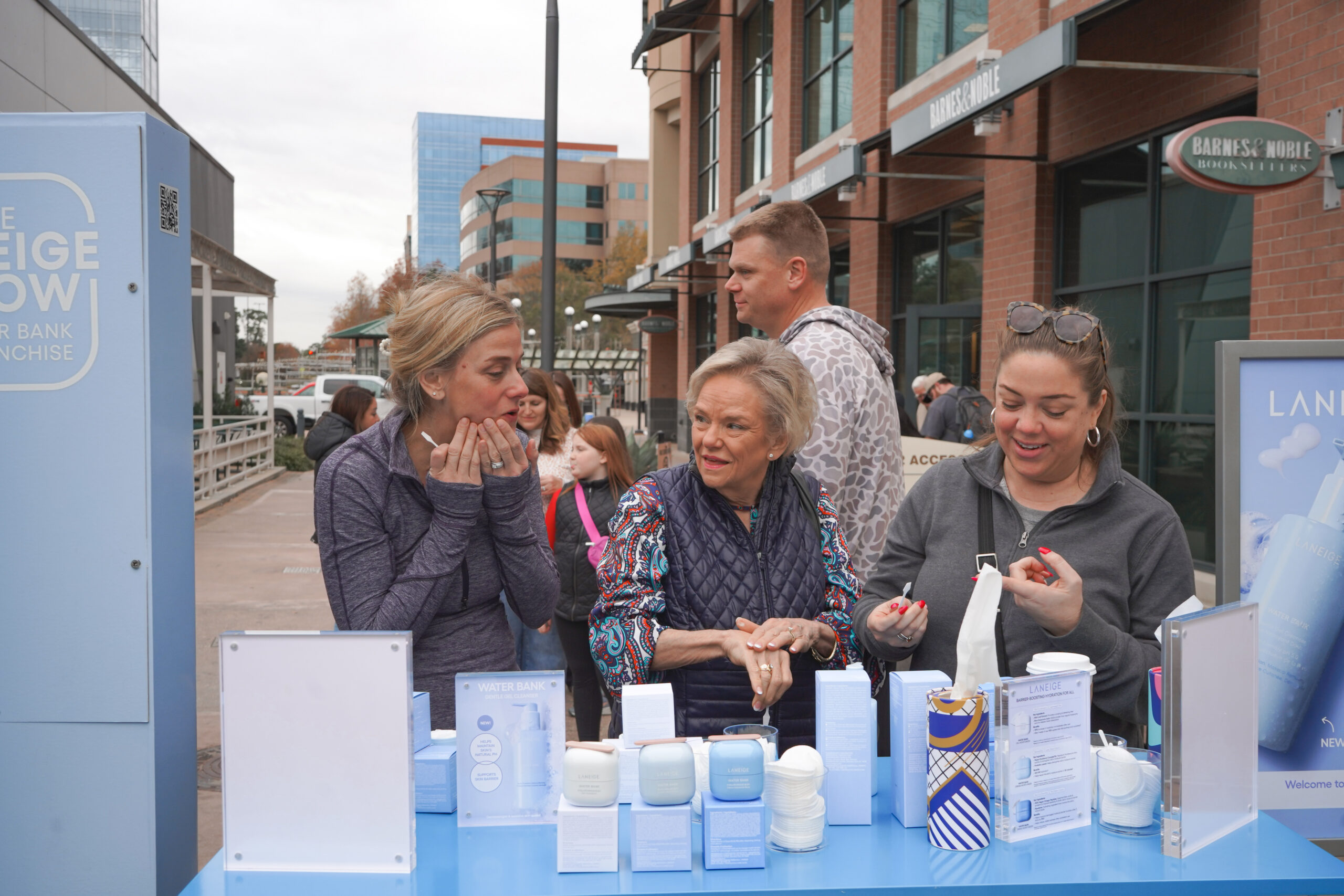 Three women talk and look at skincare items at an outdoor booth, with a line nearby.