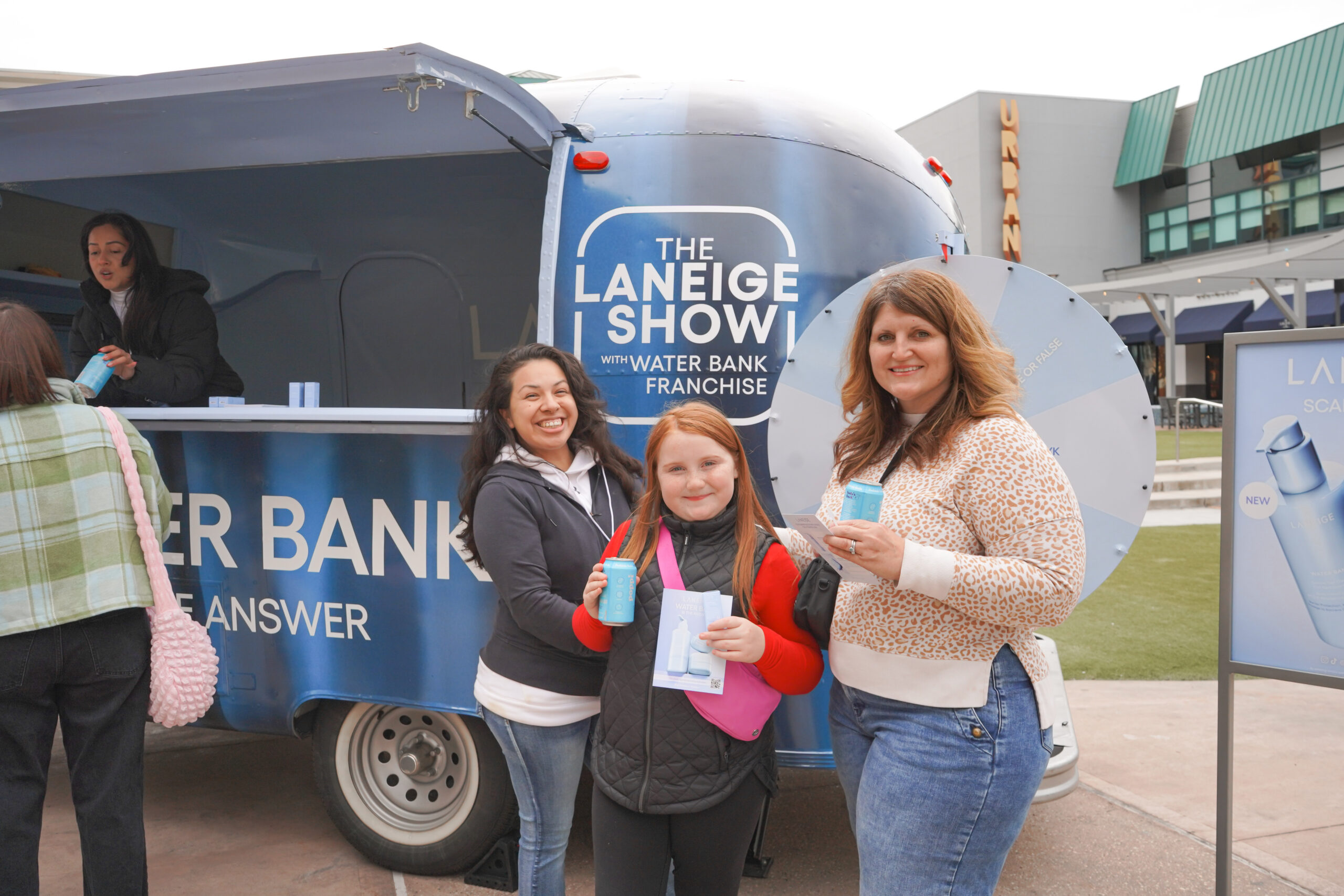 Three people hold products and smile in front of a blue Laneige trailer at an outdoor event.