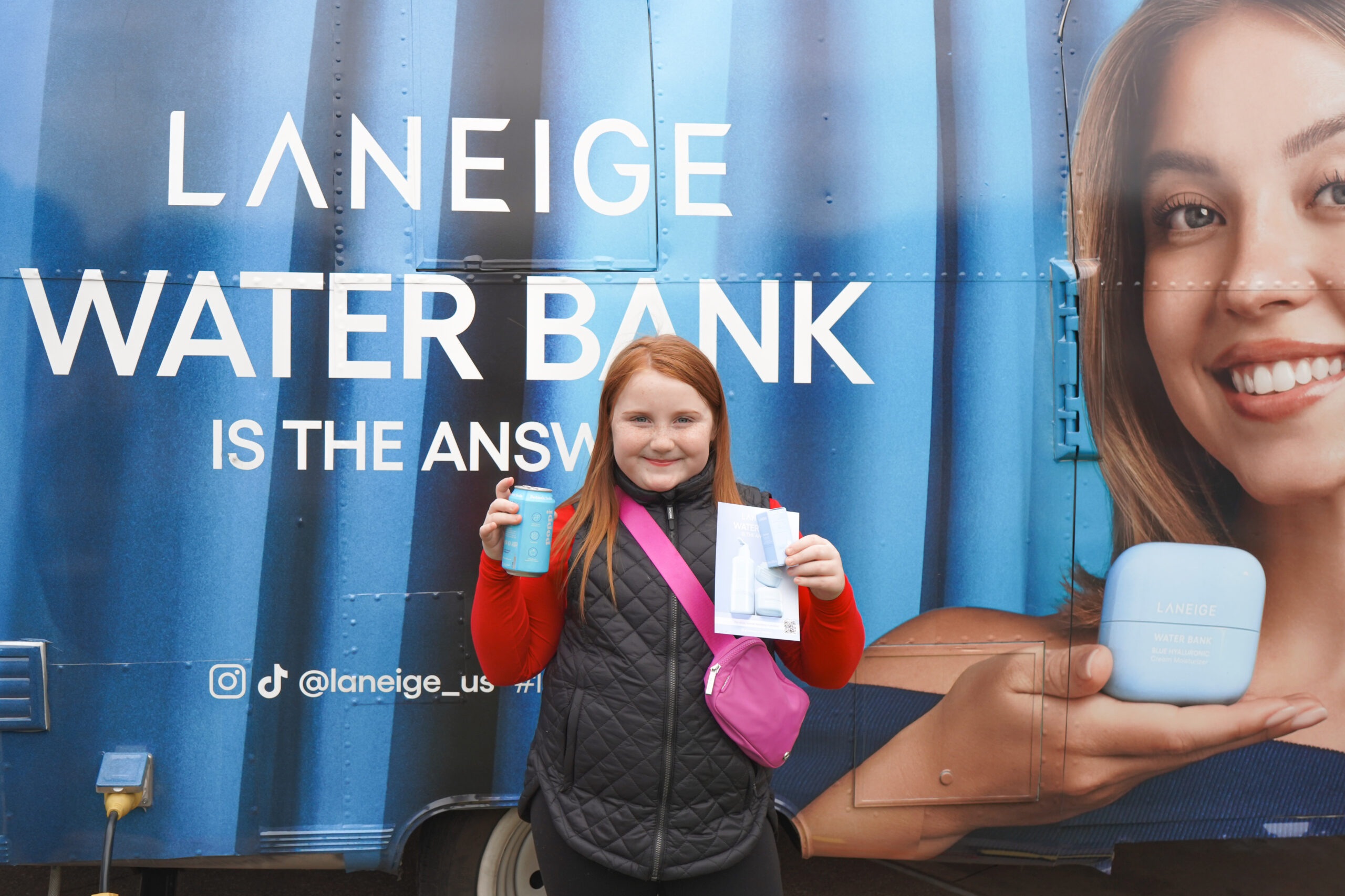 Person with red hair holds a blue cup and brochure by a LANEIGE Water Bank truck display.