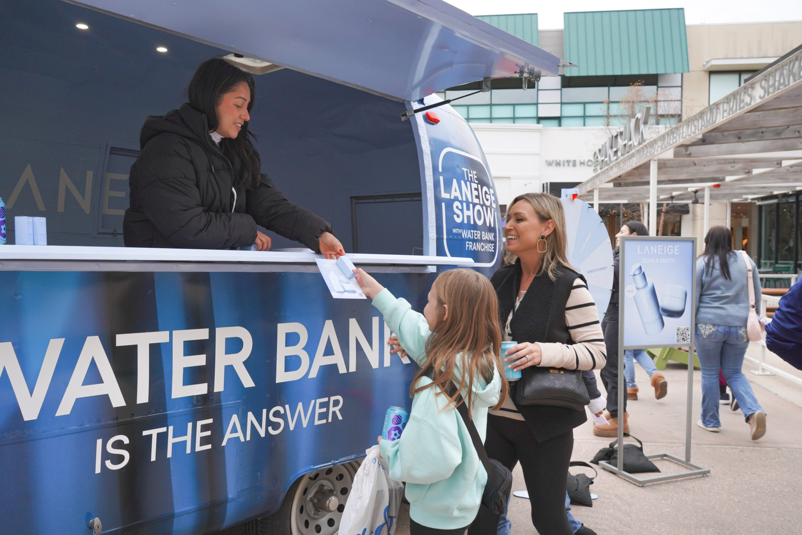 A woman and girl receive a sample from an attendant at a blue Laneige Water Bank truck outside.