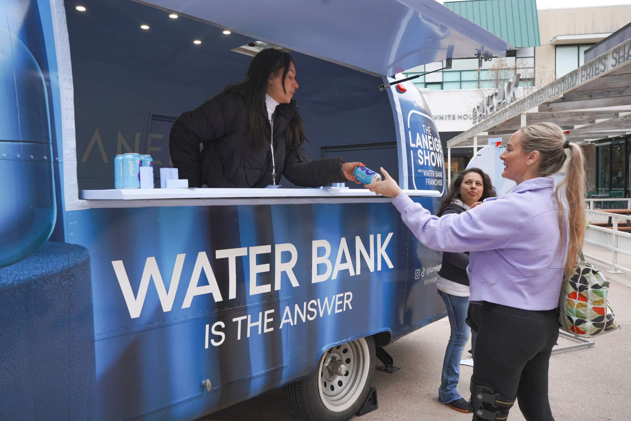 A woman gives bottled water to another woman from a blue WATER BANK IS THE ANSWER truck outside.