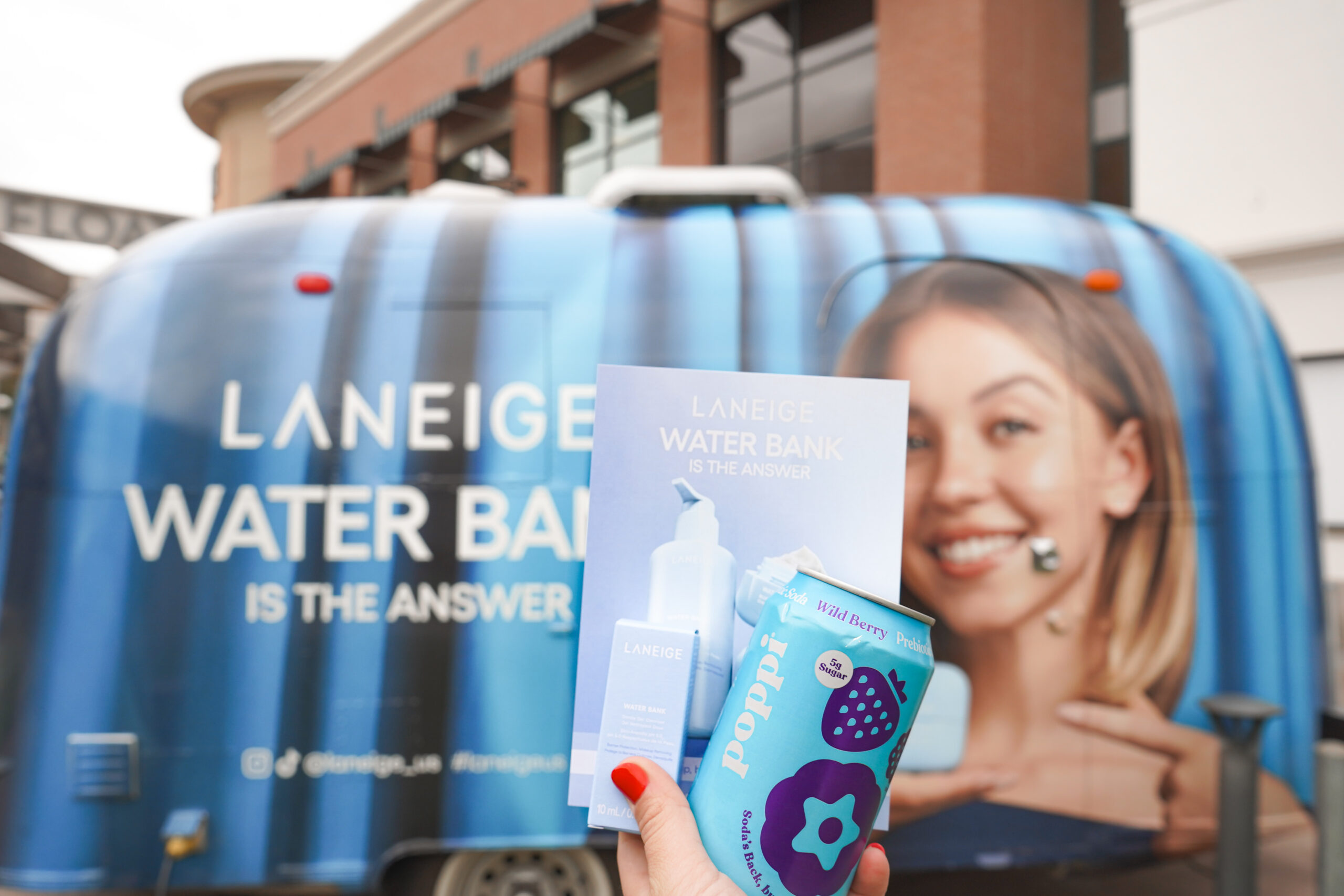 Hand holding Laneige pamphlet and blue Poppi can in front of branded Laneige van with woman's face.
