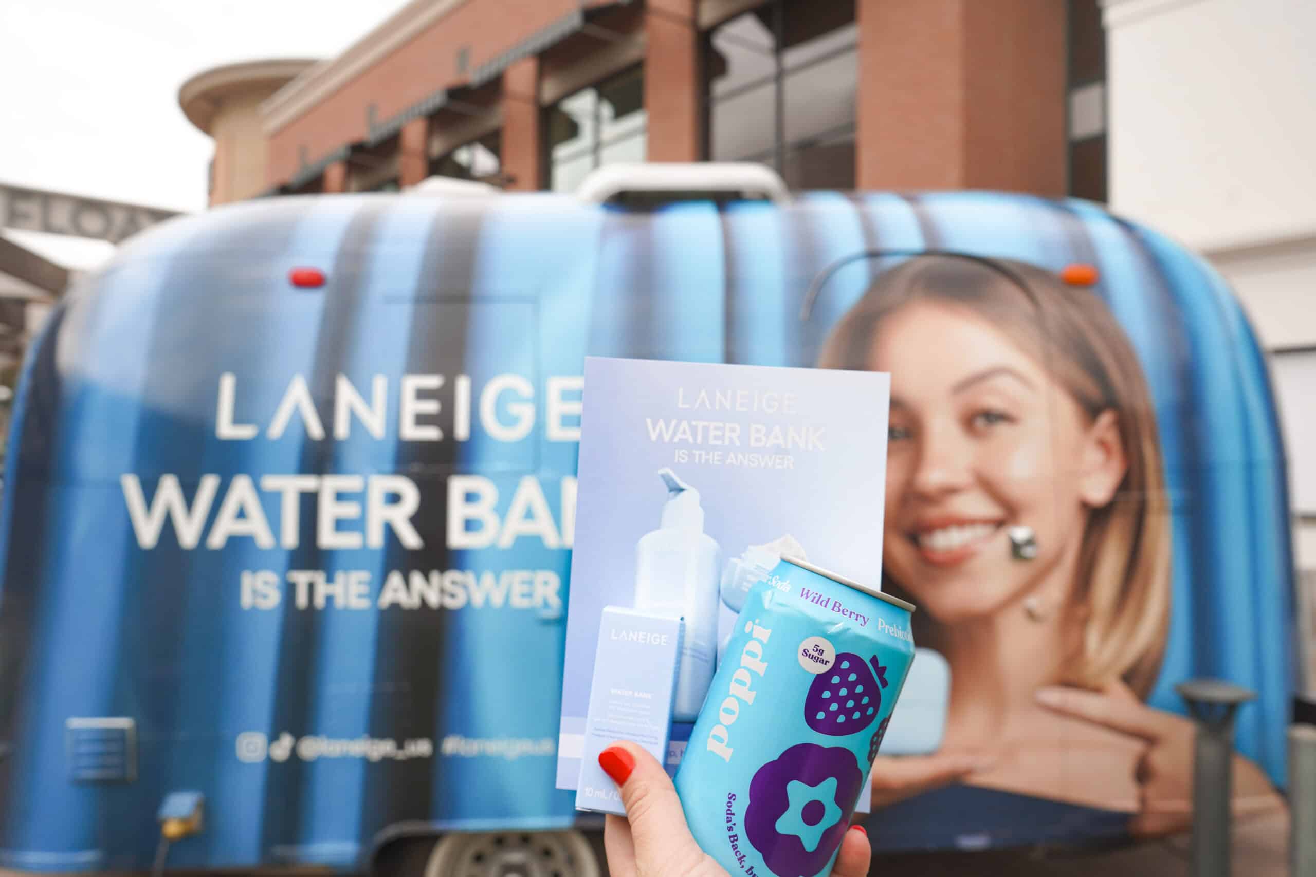 Hand holding Laneige pamphlet and Poppi can in front of trailer with Laneige ad showing a woman.