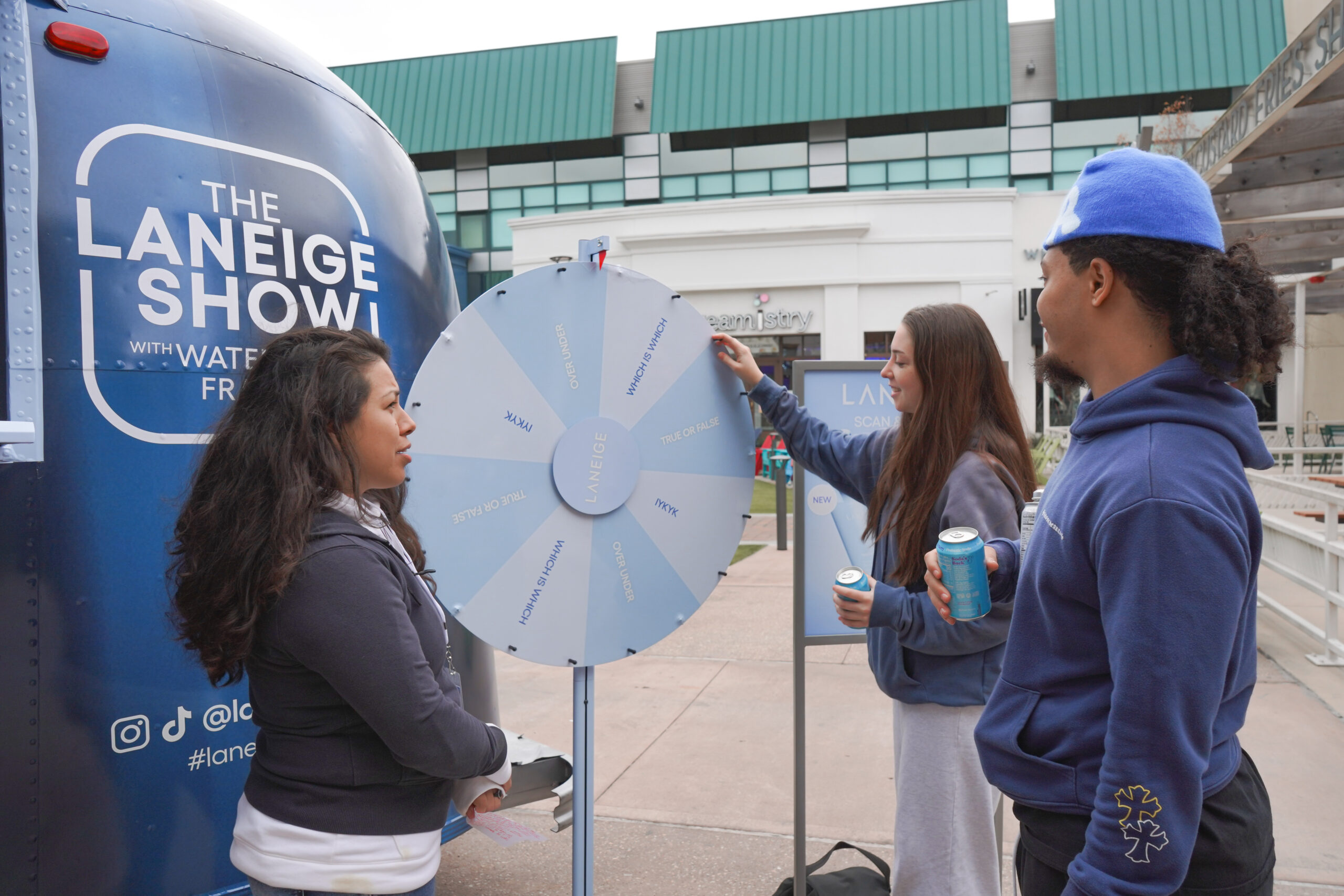 Three people by a spinning Laneige Show prize wheel; one spins, two watch holding canned drinks.
