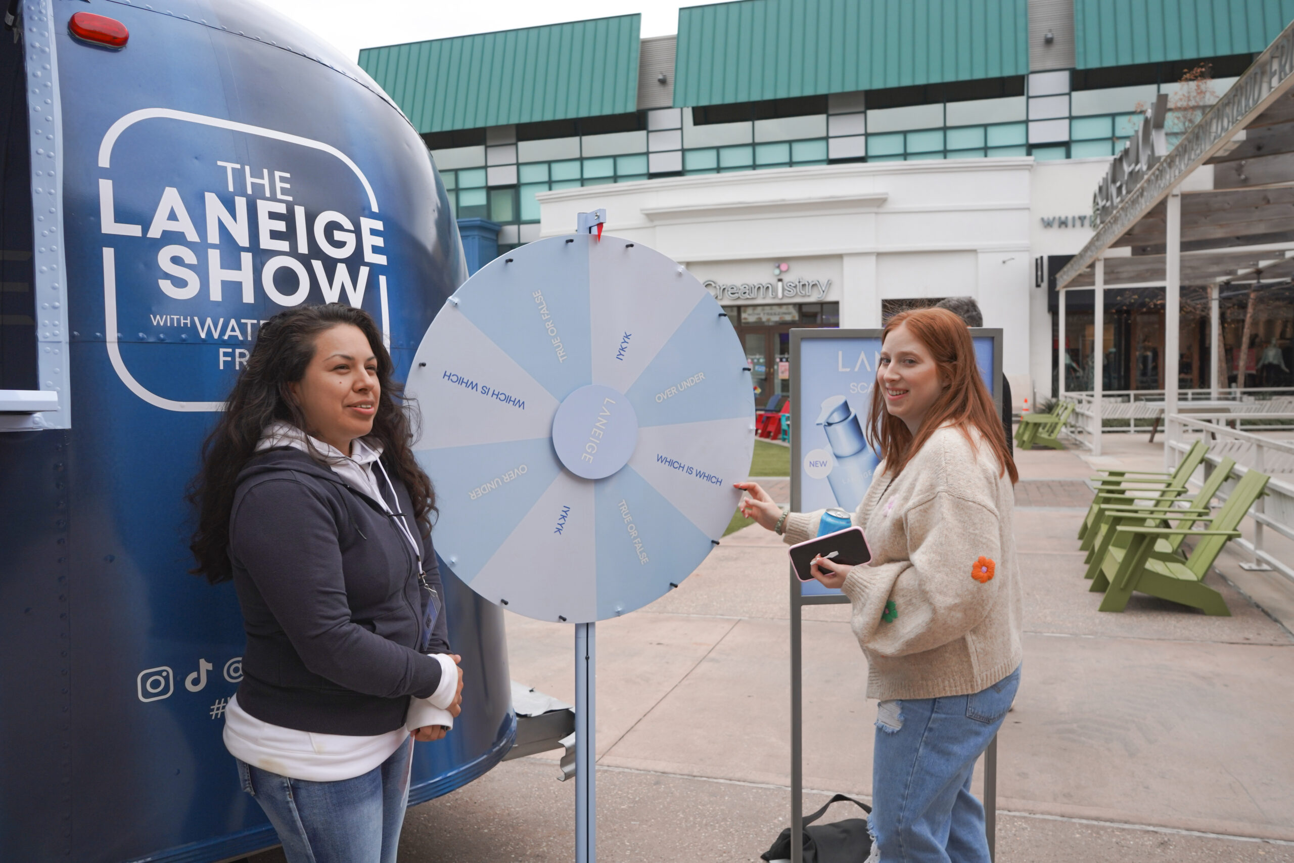 Two women by a blue prize wheel at a Laneige booth; one spins while the other watches, both smiling.