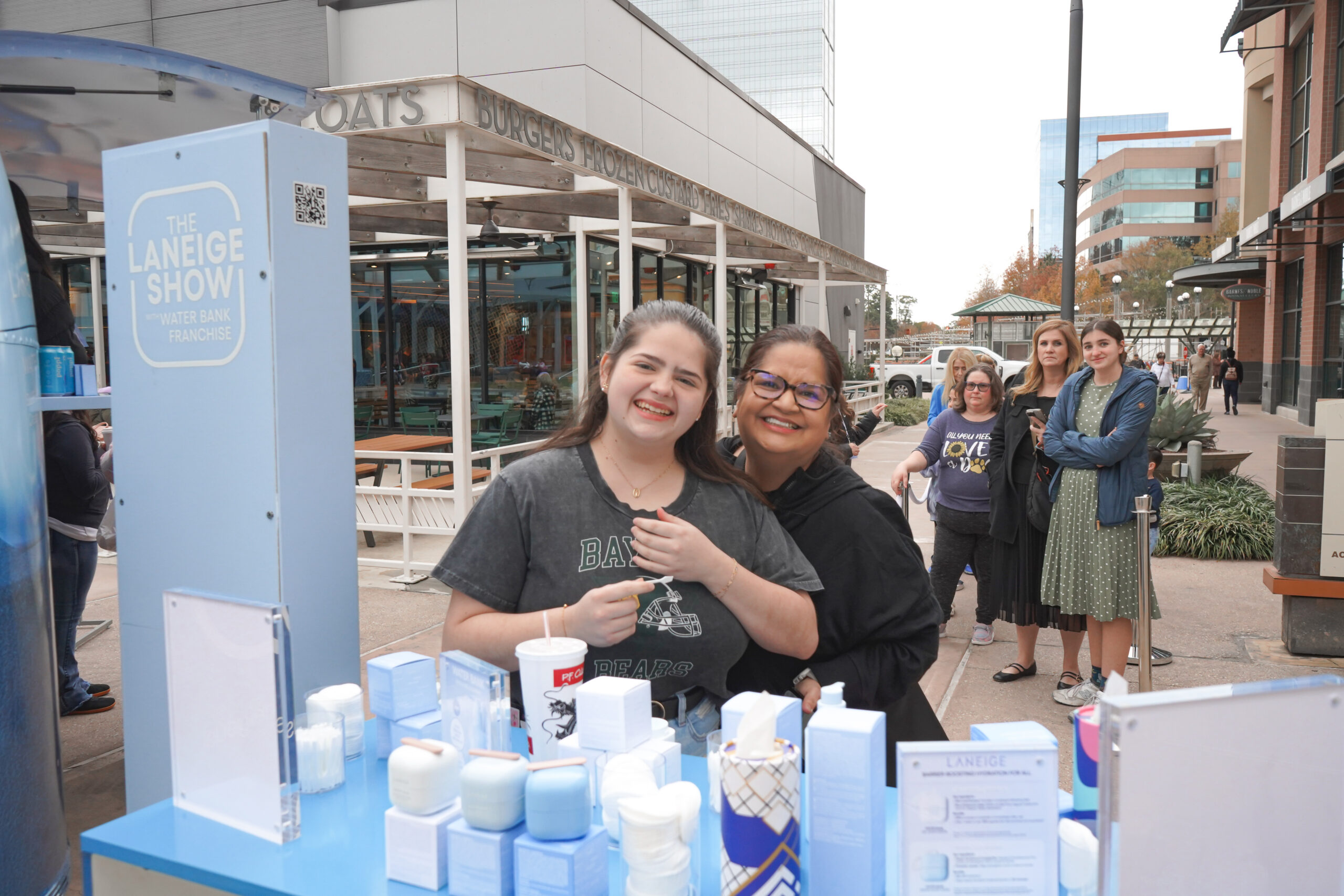 Two women hold Laneige skincare products at an outdoor booth, with a line and city buildings behind.