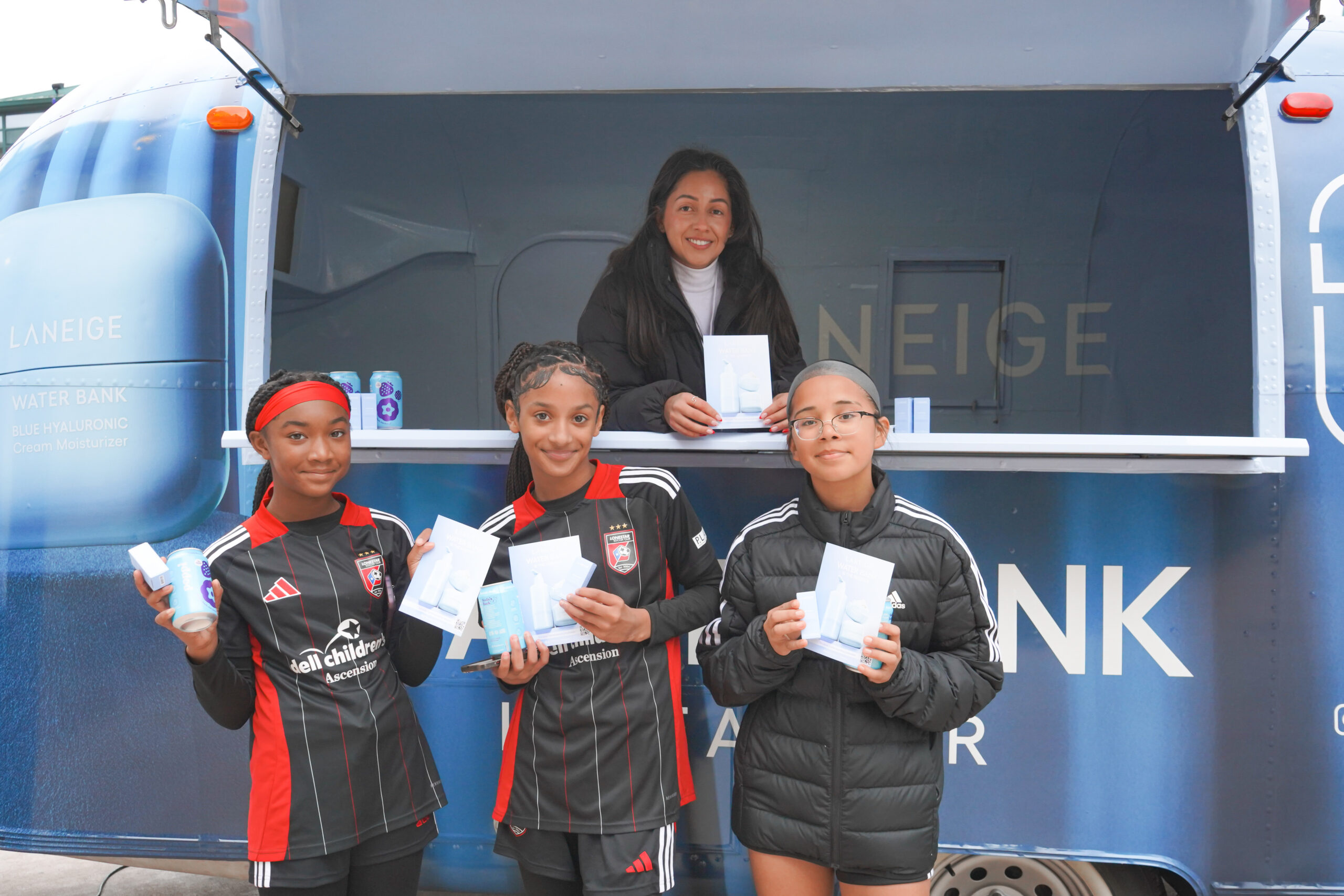 Three girls in soccer uniforms and a woman hold Laneige products by a blue Laneige Water Bank truck.