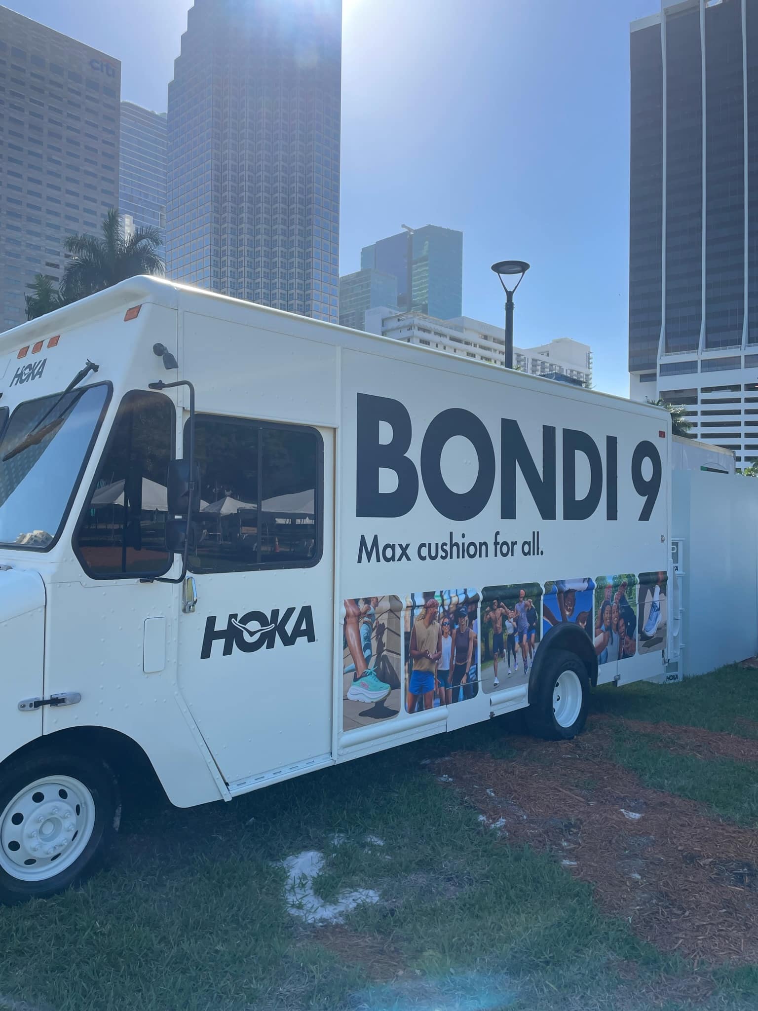 White truck with HOKA “BONDI 9 Max cushion for all” branding parked on grass by tall city buildings.