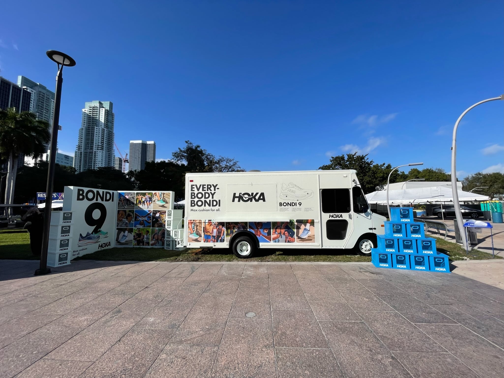White HOKA van with “EVERY BODY BONDI” and “BONDI 9” parked by blue cubes, city and palms behind.