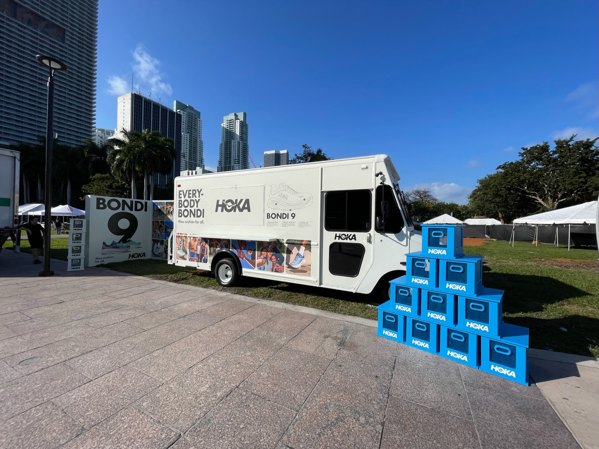 White Hoka truck by stacked blue boxes and Bondi 9 sign, with buildings and palm trees nearby.