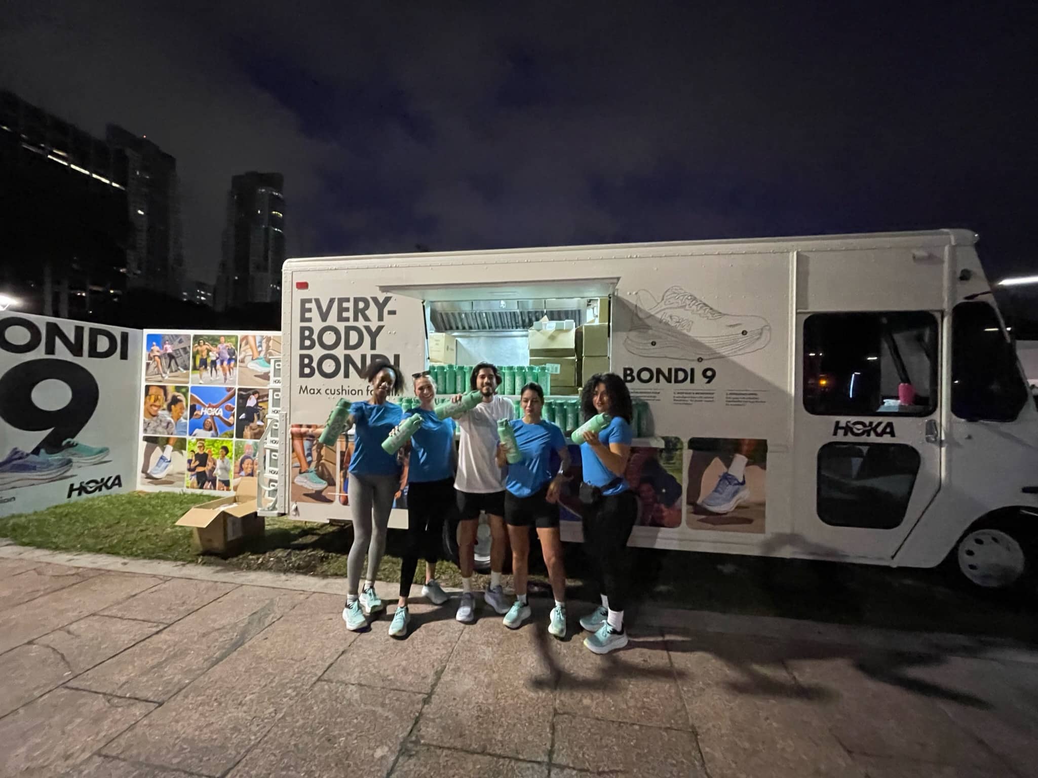Five people smiling in front of a Hoka BONDI 9 food truck at night, display board to the left.