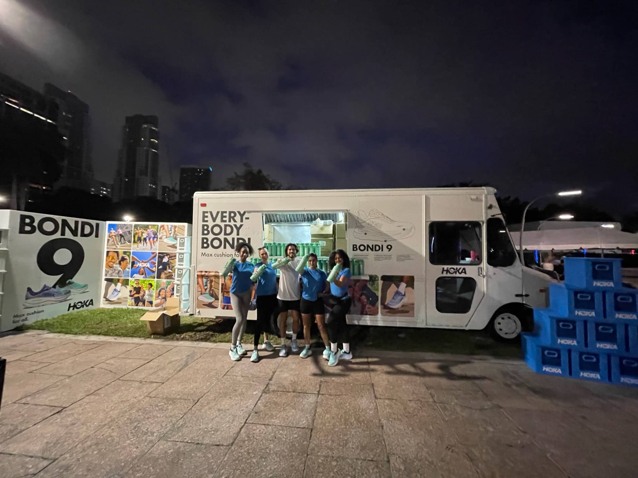 Five people in blue shirts stand by a white HOKA BONDI 9 van at night with city buildings behind.