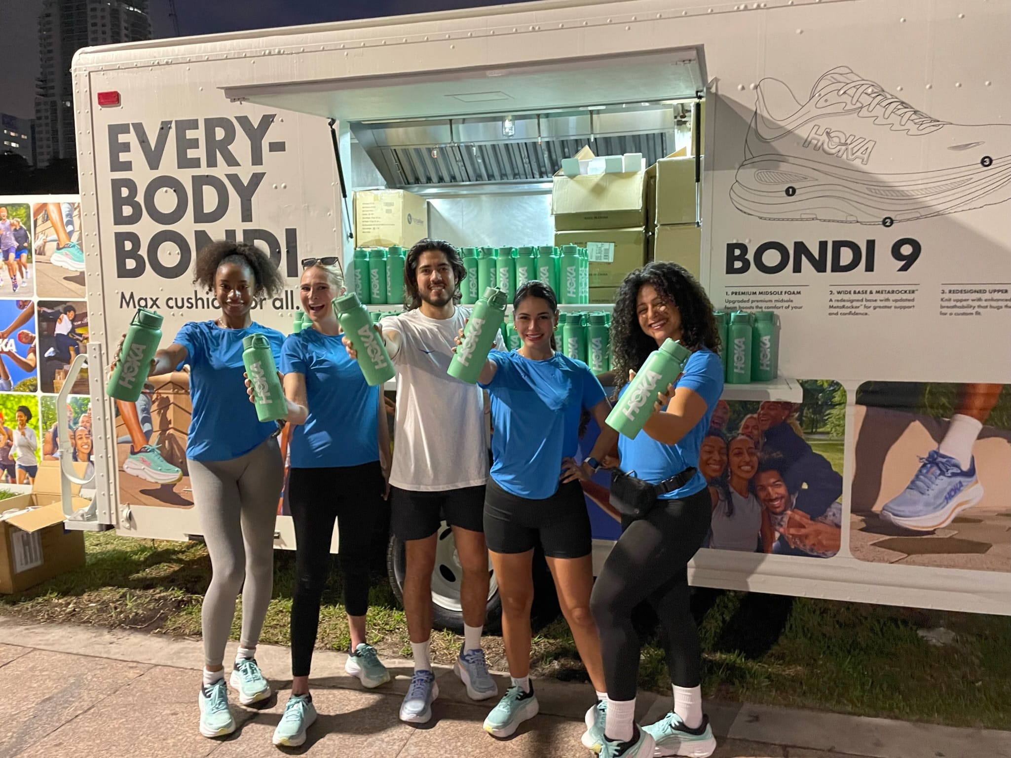 Five people with mint green water bottles stand by a Hoka truck showing sneakers and its slogan.