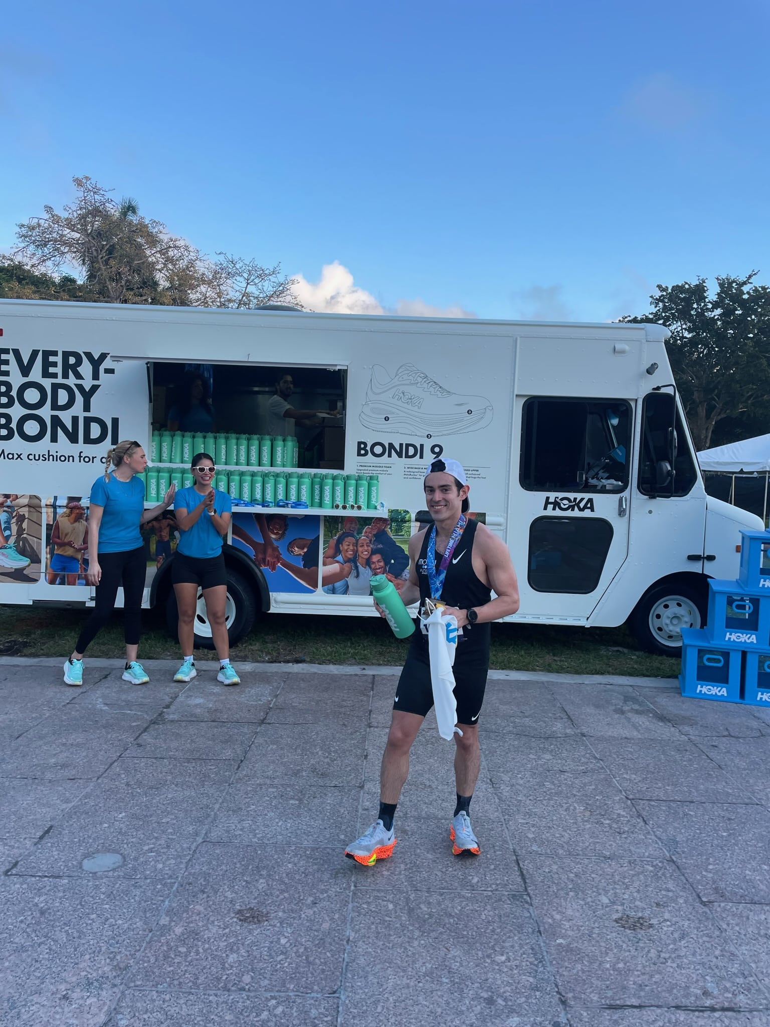 Runner with water bottle and towel smiles by a HOKA truck as two women in blue applaud nearby.