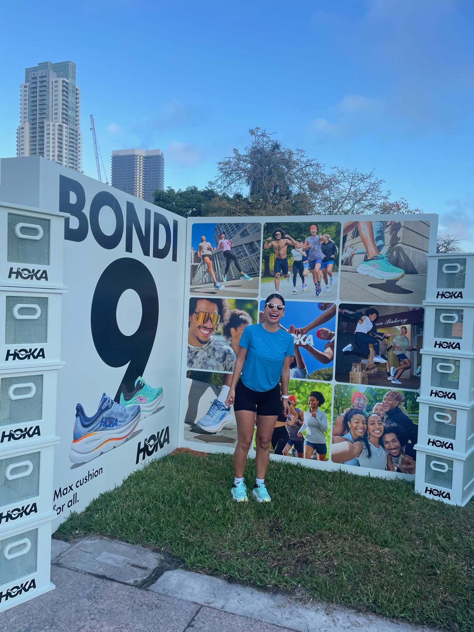 Woman in athletic wear smiles by a Hoka shoe display wall with "BONDI 9"; city skyline behind.