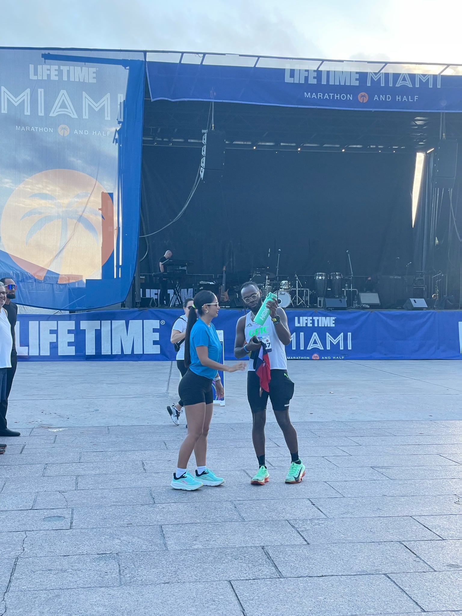 Runner with medal and towel stands smiling with a woman near Miami Marathon stage and instruments.