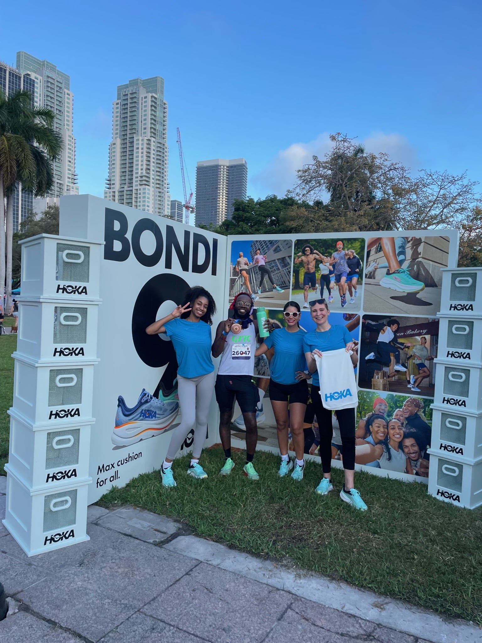 Four people smile in front of a BONDI 9 Hoka display with shoe boxes, buildings, and trees behind.