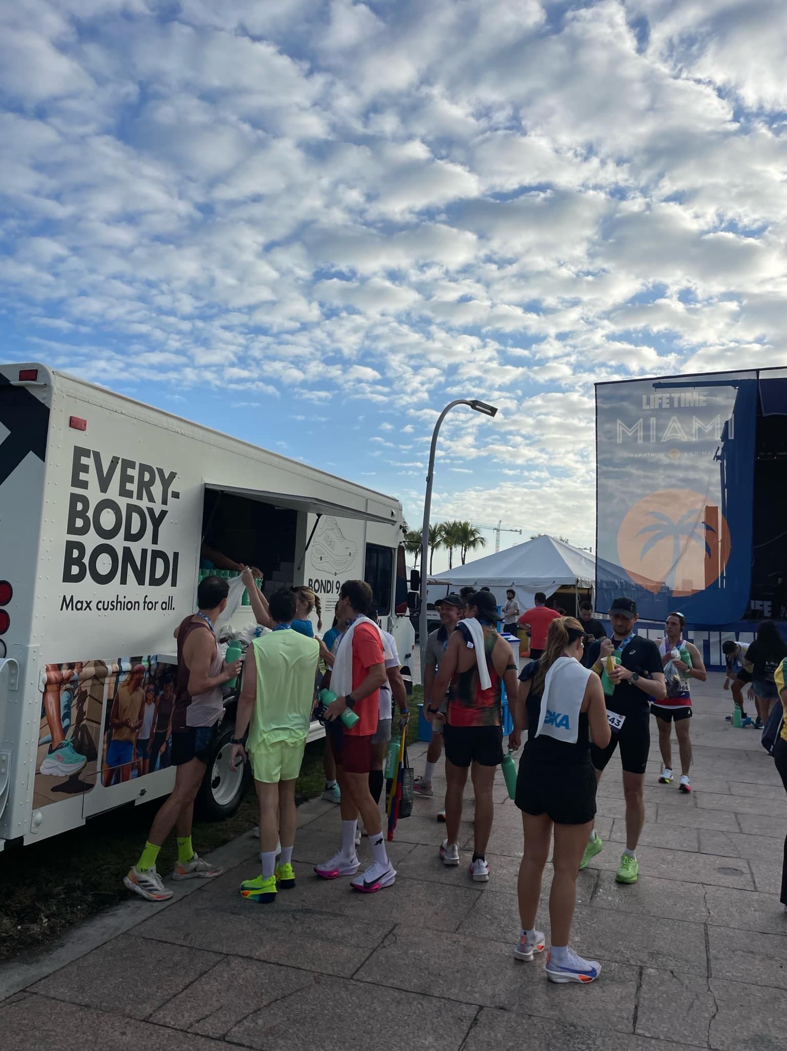Runners stand near a white EVERYBODY BONDI truck, with a MIAMI sign and palm trees behind them.