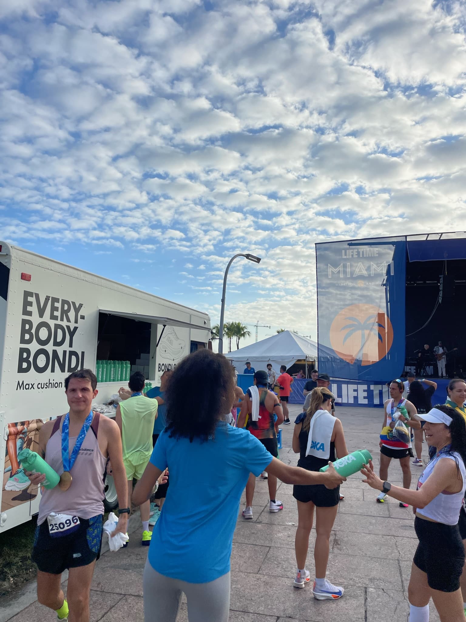 A woman gives water to marathon runners near a stage and “EVERY BODY BONDI” truck; some wear medals.