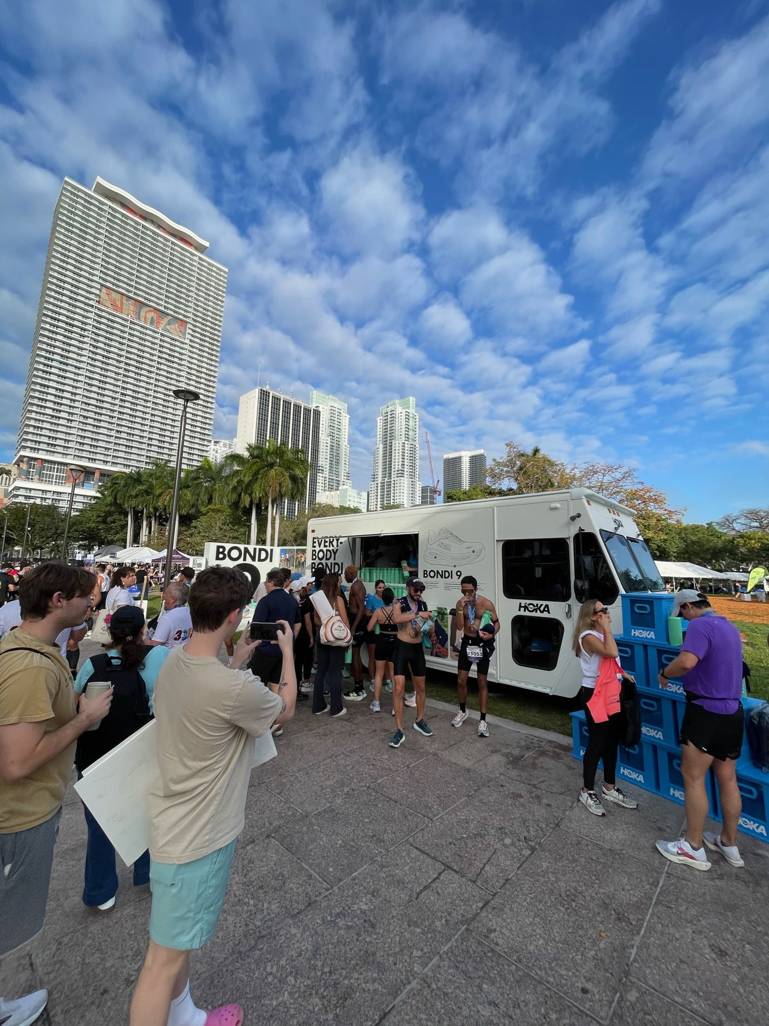 People gather near a branded van in a city plaza; blue shoe boxes are stacked beside the van.