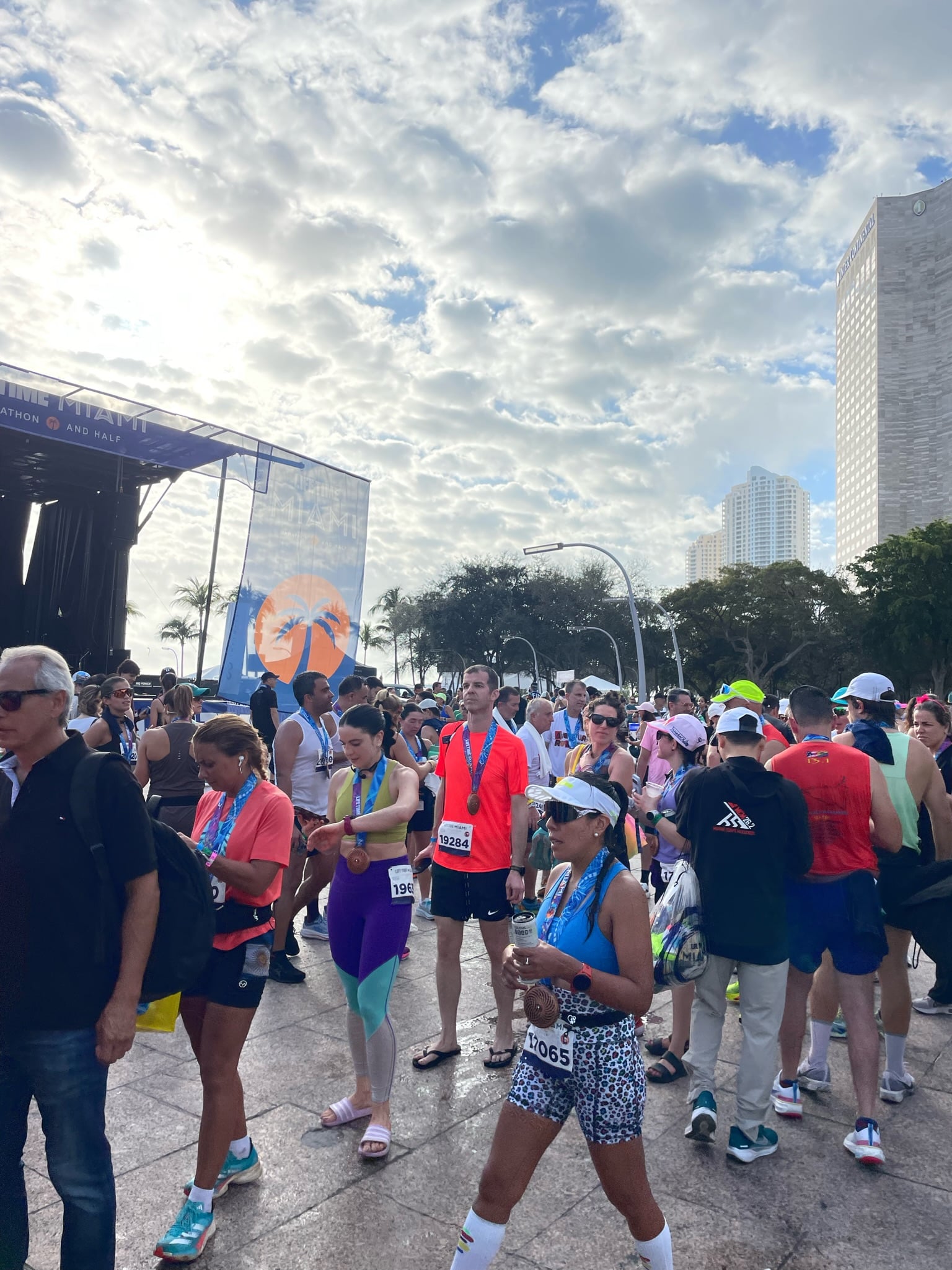 Marathon runners with medals and race bibs gather near a stage outdoors; some use phones.