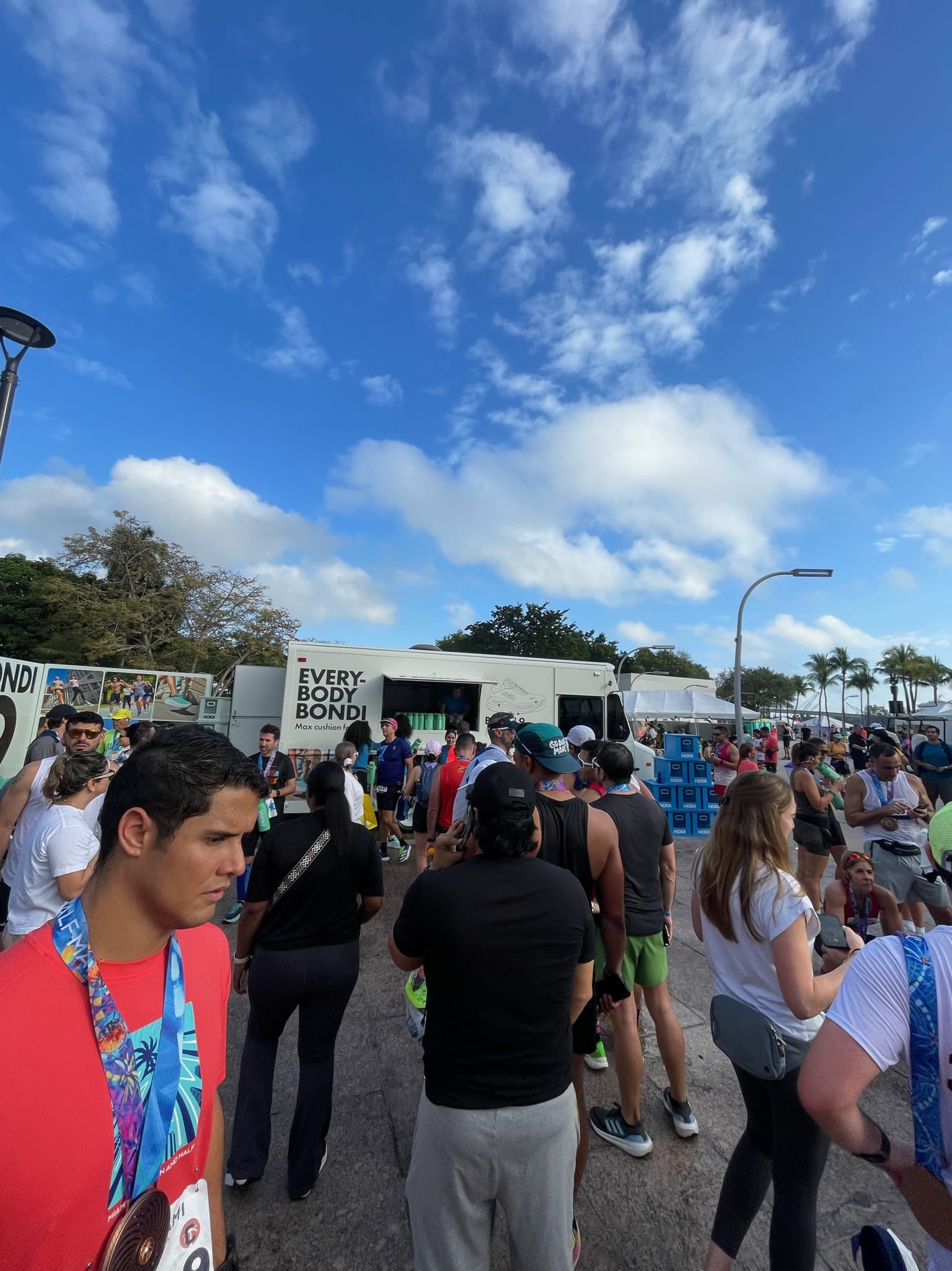 People, some with medals, stand by a white "EVERYBODY BONDI" truck near palm trees and blue bins.
