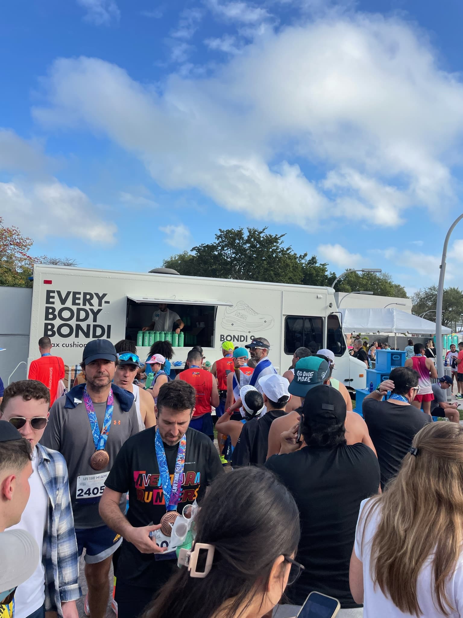 Marathon runners with medals and bibs line up at a white food truck, trees and tents behind.