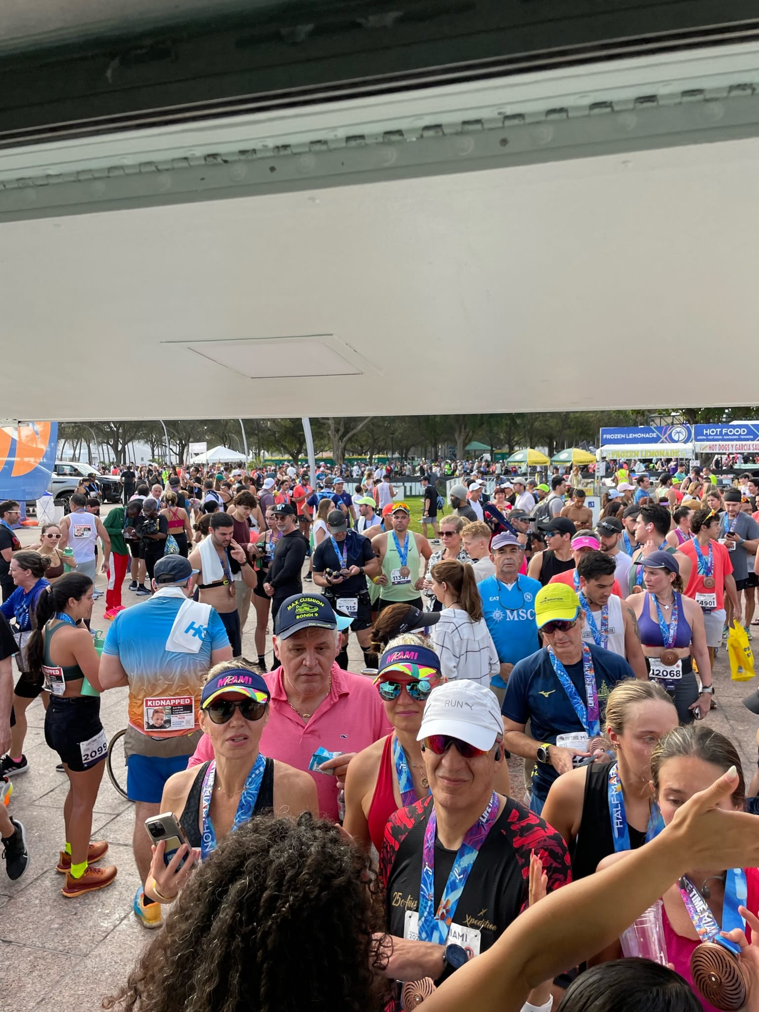 Marathon runners with medals gather near a finish line, tents and vendors in the background.